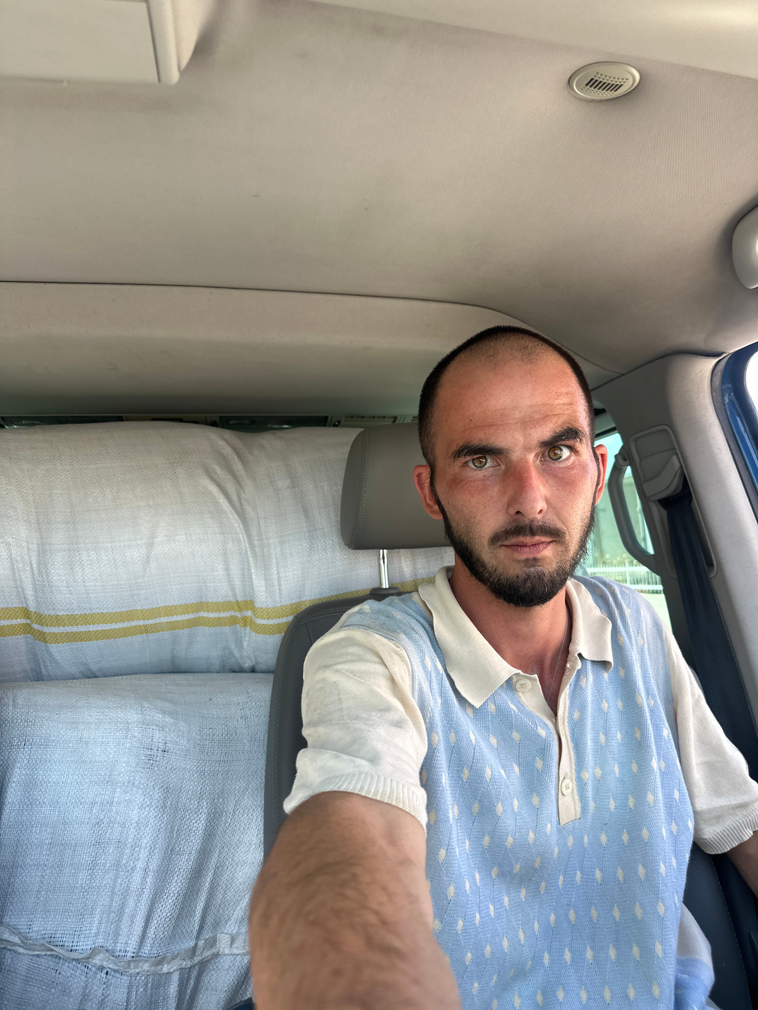 A man with short brown hair and a beard is sitting in the driver's seat of a vehicle. He is wearing a light-colored polo shirt with a blue diamond pattern. The interior of the vehicle is visible, including the headrest of the passenger seat and what appears to be large, white, woven bags stacked behind the seats. A ventilation grille is visible on the ceiling of the vehicle. The image appears to be taken in Rakića Kuće, Montenegro, based on the context provided.