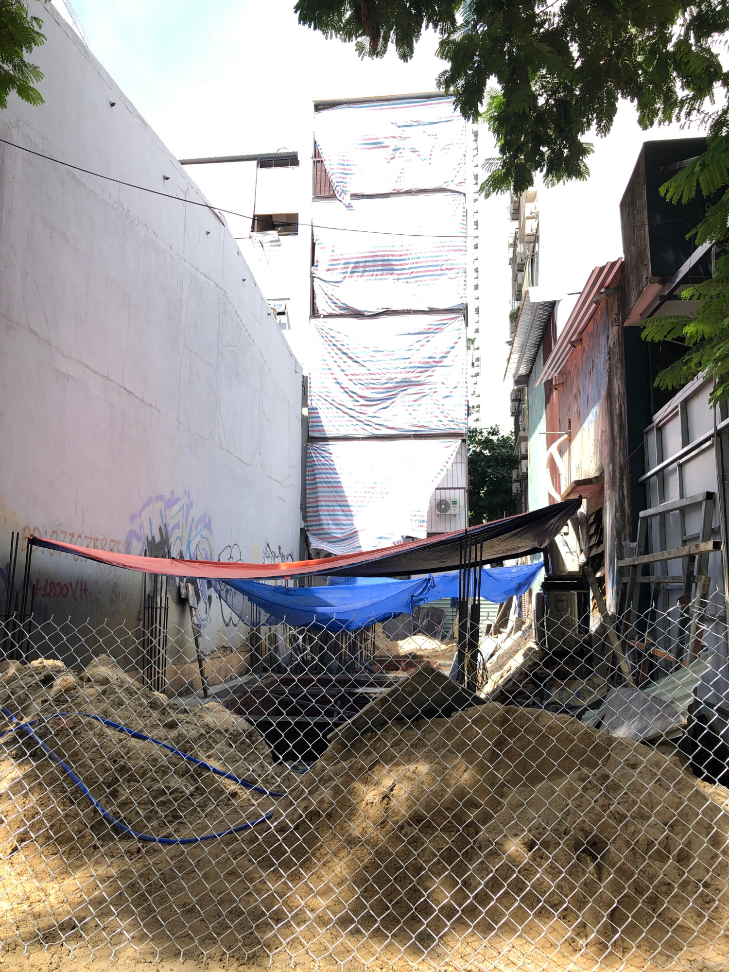 This image captures a construction site in Đà Nẵng, Vietnam. The scene is oriented vertically, showing a construction site in progress. In the foreground, a chain-link fence with a diamond pattern encloses a pile of sandy soil, some of which appears to have been excavated. Blue and orange tarps are draped over something within the construction area, partially obscuring the view. Behind the fence, several buildings are visible. Some appear to be older, with weathered facades, while others are under construction or renovation, indicated by striped tarps covering large sections of their exterior. The sky is bright, suggesting it is daytime. The overall impression is one of urban development and construction activity in a Vietnamese city. No people are clearly visible in the image. There is some graffiti on a concrete wall in the lower right quadrant of the image, but the text is not legible.