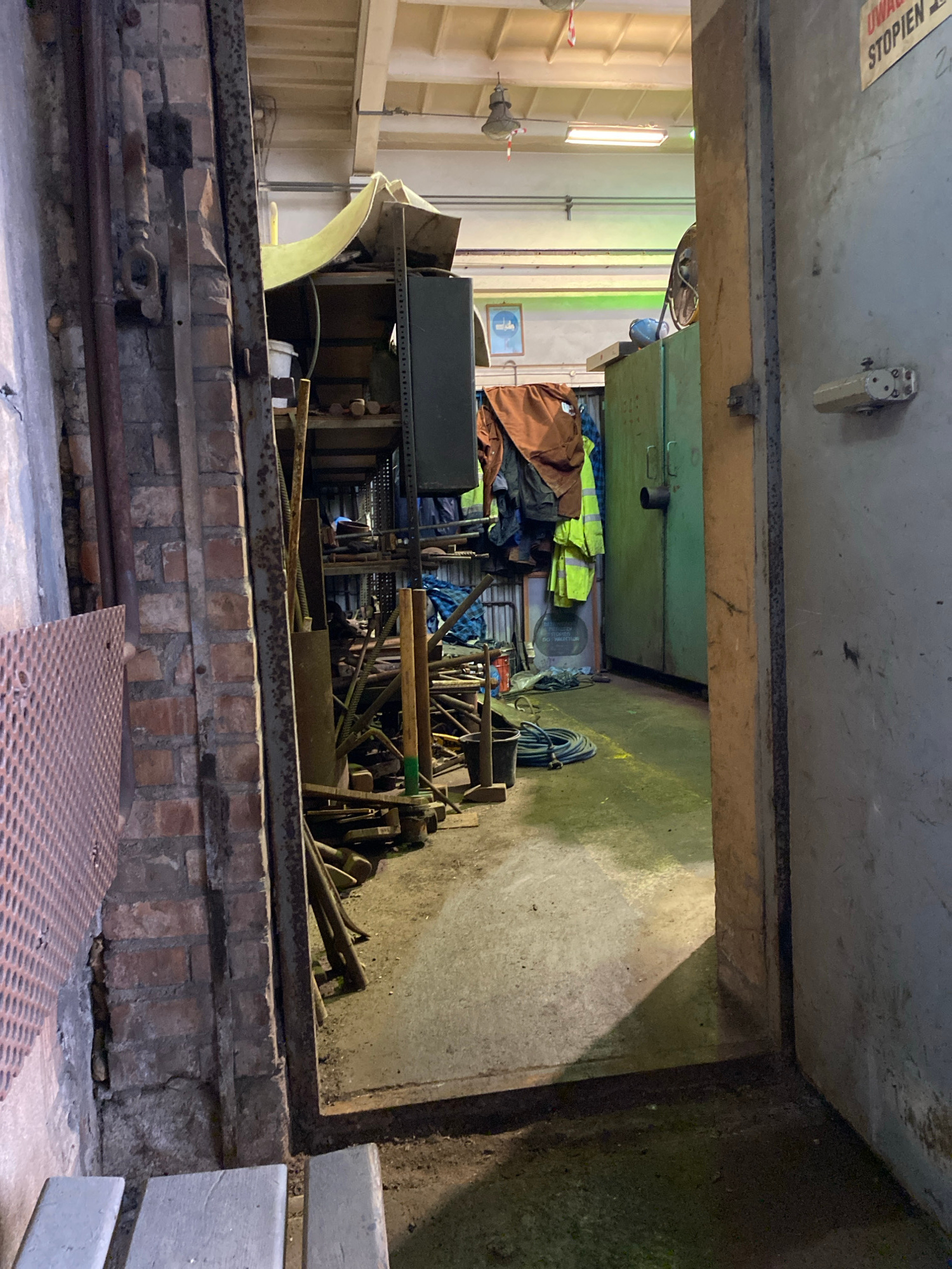 The image depicts a cluttered storage area or workshop, possibly within an industrial or maintenance facility. The perspective is from within a narrow passage looking into a larger space filled with various objects. Dominating the foreground is a brick structure with metal pipes and fixtures.
Inside the larger space, shelves are stacked with tools, equipment, and what appear to be safety vests in neon yellow and orange, along with brown workwear. A coiled blue rope is visible, as well as various metal rods and pieces of machinery. The walls of the inner space are a muted green and grey. The lighting suggests it could be daytime, as some light streams in from the left, illuminating the shelves and the stacked items. There are no people visible in the image.
On the ceiling in the upper left of the image, there is a warning sign with text in what appears to be red and white. The text reads "UNROLL" and "STOPPEN" which translates to "STOP" in German. This suggests the location might have had a German presence or is in an area where German is understood. Given the provided location context of Police, Poland, this sign could be a remnant of past use or a multilingual warning. The overall impression is one of an organized but heavily used workspace, filled with the tools and materials for maintenance or production.