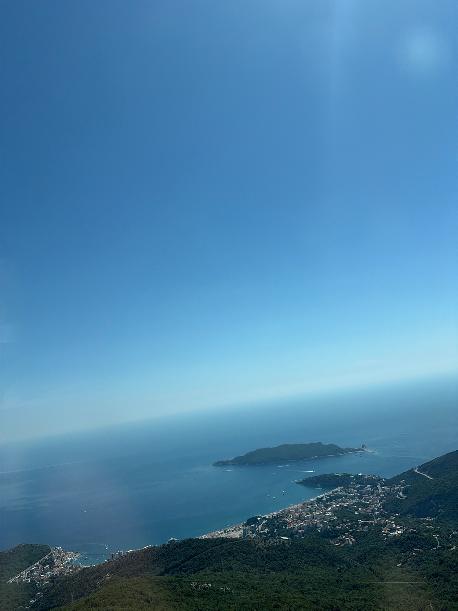 The image is an aerial view of the coast near Brajici, Montenegro, on a clear, sunny day. The vast expanse of the Adriatic Sea dominates the upper two-thirds of the frame, its surface calm and reflecting the bright blue sky. A thin band of hazy light appears at the horizon, a common atmospheric effect in aerial photography.
In the lower portion of the image, a densely populated coastal town is nestled between the mountains and the sea. Buildings, ranging in size and architectural style, are clustered together, with some appearing to be hotels or apartment complexes due to their scale and proximity to the beach. Roads wind through the town and up the steep, verdant hillsides that frame the settlement. The mountains are covered in thick, dark green foliage, indicating a lush natural environment.
To the left, a small harbor is visible with several boats docked and others in motion within the sheltered waters. Further out in the sea, to the right of the main town, a distinct island with a rocky outcrop is visible. Small boats are scattered across the water, suggesting recreational or commercial activity. The overall scene depicts a picturesque coastal resort town situated in a dramatic natural landscape.