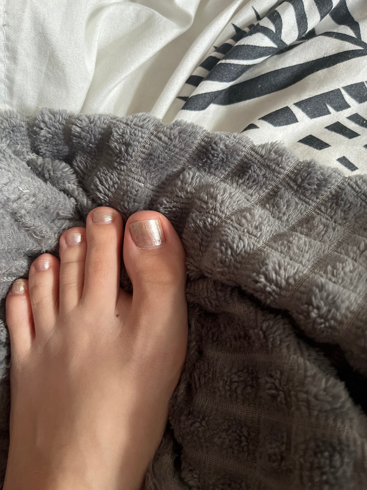 A close-up, overhead shot shows a left foot with light-colored toenail polish, resting on a textured grey blanket. Behind the blanket, a white fabric with black stripes is visible. The foot is positioned in the center of the frame, with the toes angled slightly towards the right. The lighting is soft, casting subtle shadows. No specific location within Volgodonsk, Russia is identifiable.