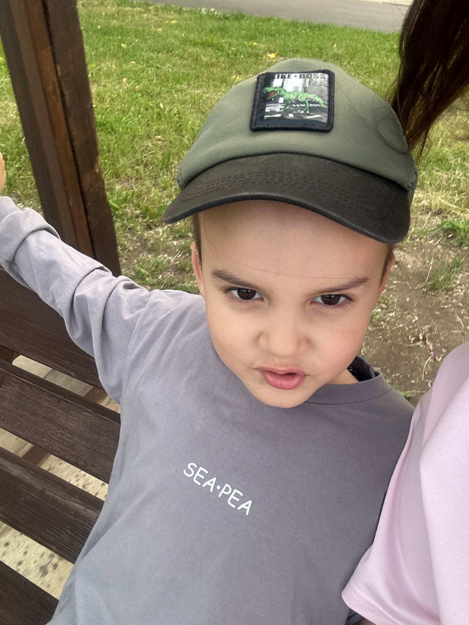 A young boy, approximately five years old, is captured in a close-up, high-angle selfie. He is wearing a grey long-sleeved shirt with the text "SEA-PEA" printed in white, and a dark green baseball cap with a patch depicting a dinosaur and the words "LIKE BOSS". The boy has dark brown eyes and is looking directly at the camera with a slightly puckered mouth. He is sitting on a wooden bench, partially obscured by a dark brown wooden structure. The background consists of green grass and dirt, suggesting an outdoor environment. Another person, whose shoulder and arm in a pink garment are visible on the right side of the frame, is also present. The lighting indicates it is daytime. The scene appears to be in a park or outdoor recreational area within the city of Lytkarino, Russia.