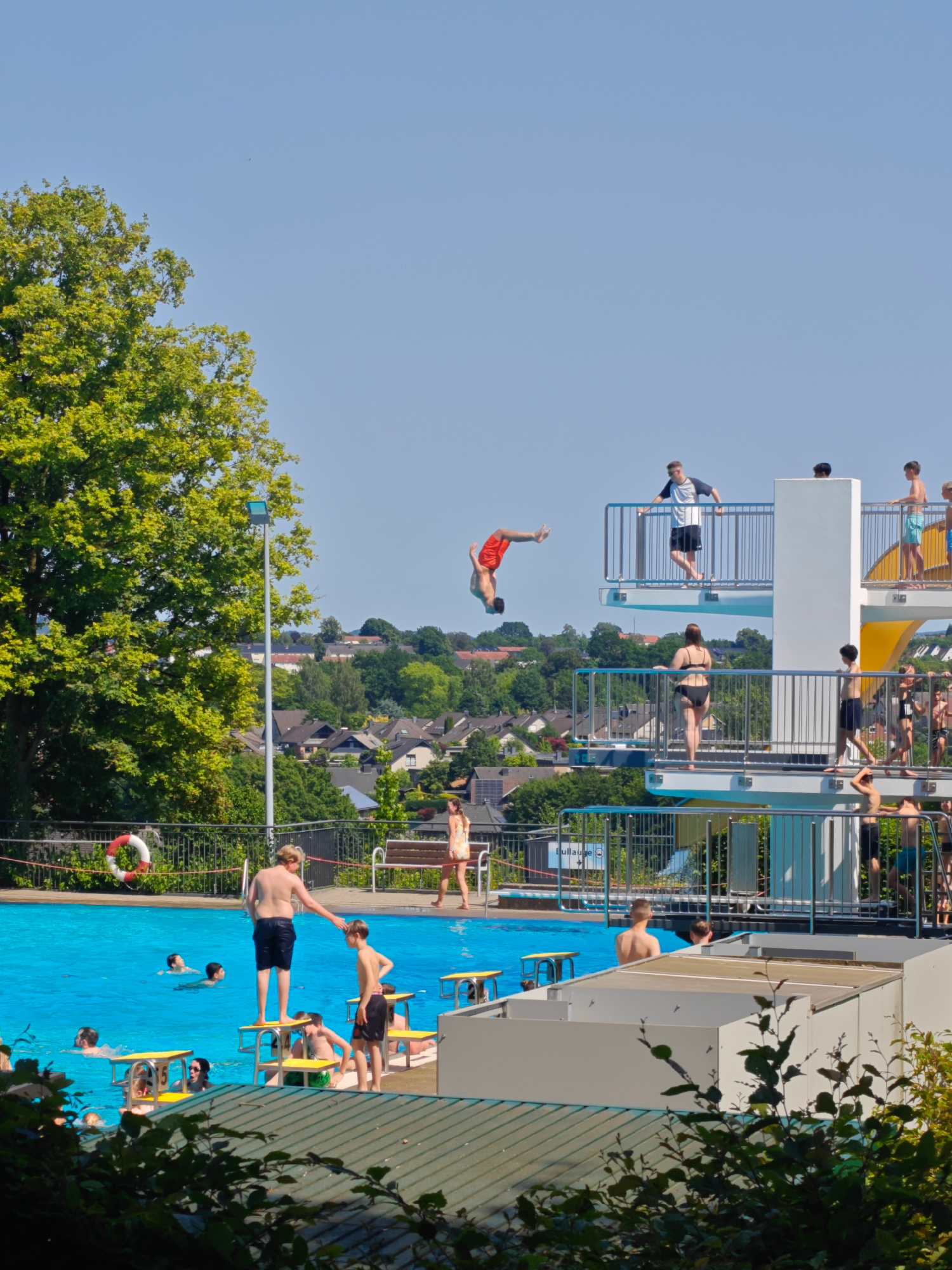 This media file depicts a bustling outdoor swimming pool on a clear, sunny day, likely in Georgsmarienhütte, Germany, given the location context. The scene is filled with people enjoying the water and the diving platforms.
The primary subjects are people of various ages engaged in swimming and preparing to dive. A notable activity is a young man in mid-air performing a flip off a high diving board, showcasing a moment of athletic prowess. Several individuals are also seen standing on the diving platforms, either waiting their turn or observing the divers. Others are in the pool, swimming or standing in the shallow end. Objects present include the diving platforms, railings, yellow diving boards, and a life preserver ring with red accents hanging on a fence. The setting is an open-air swimming facility with a bright blue pool and lush green trees and residential buildings in the background.
The time of day appears to be midday, suggested by the bright sunlight and clear sky, indicative of good weather suitable for outdoor recreation. The overall mood conveyed is one of energetic leisure and summer fun. The visible text "pullauge" with a circular logo is present on a blue section of the pool edge.