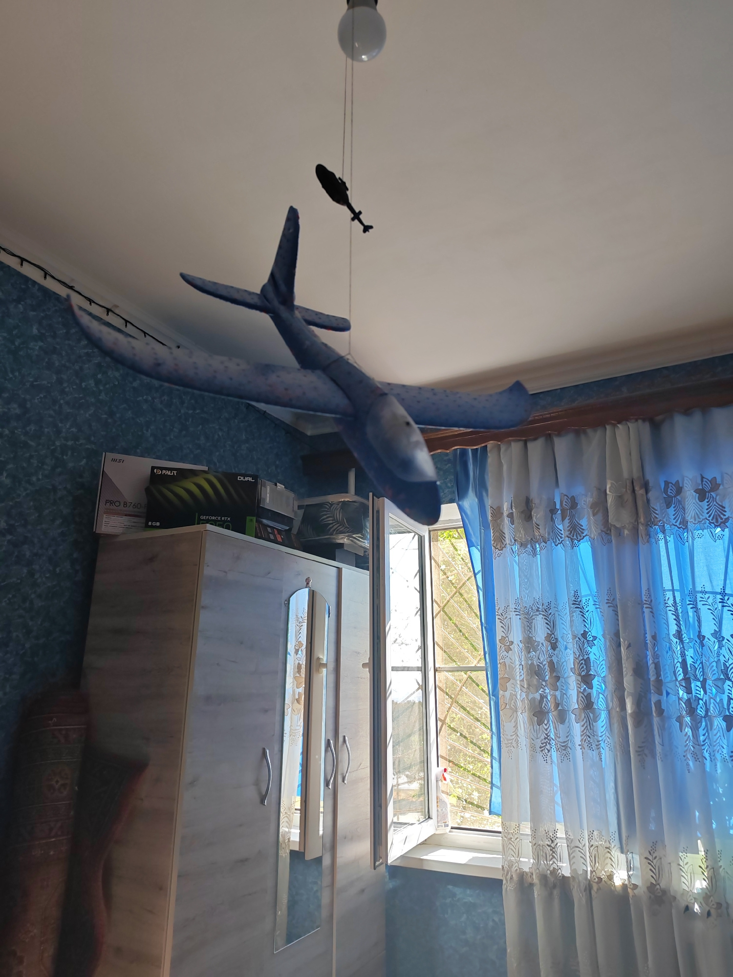 A ceiling-mounted airplane model and a smaller helicopter model hang from the ceiling in a room. Below them, on top of a light-colored wardrobe, are several computer component boxes, including a Palit GeForce RTX 3050 graphics card box. To the right of the wardrobe is a window with blue and white patterned curtains. The room appears to be a bedroom or living space. The location is not specified in the image.