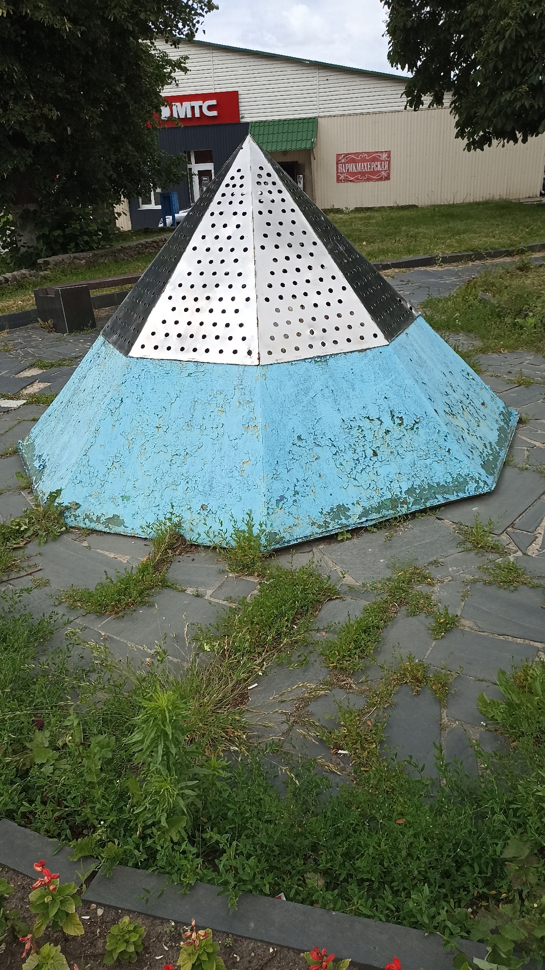 The image shows a pyramid-shaped structure with a silver, perforated upper section and a blue, peeling painted lower section. The structure is located outdoors on a paved area with scattered grass and weeds growing between the paving stones. In the background, there is a building with a white, horizontally paneled facade. A red sign with the text "MTC" is visible on the building, and another red sign with Cyrillic text, likely "ПАРИКМАХЕРСКАЯ" (Hairdresser's), is also present. Trees and foliage are visible to the left and right of the building, and part of a park bench is to the left of the pyramid. The scene appears to be in a public space, possibly a town square or park. The location is identified as Kapyĺ, Belarus.
