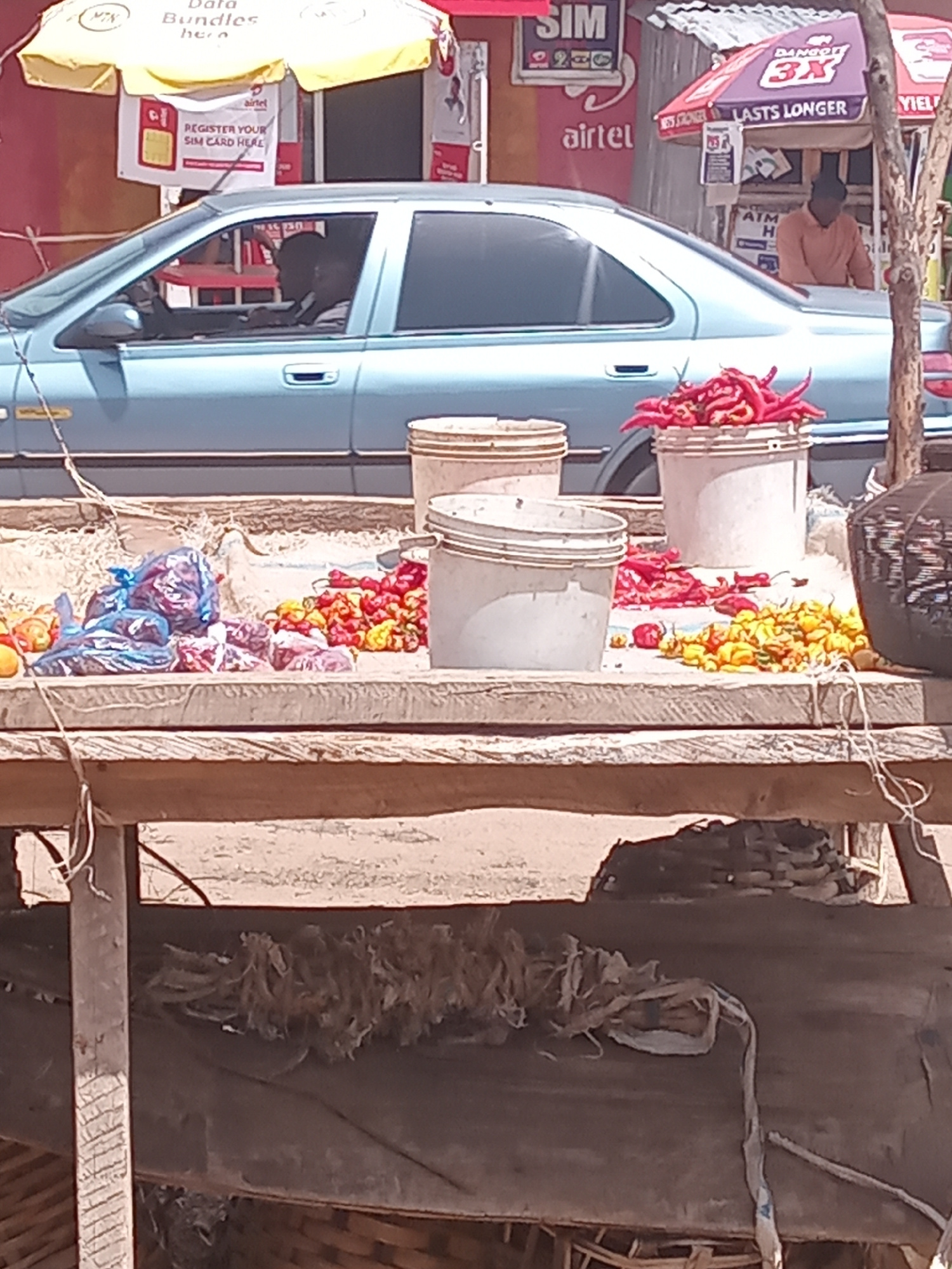 This is a medium-shot, vertically oriented photograph taken outdoors in what appears to be a bustling marketplace or roadside vendor area. The main subject in the foreground is a light blue car, partially visible. To the left of the car, a yellow umbrella is set up, advertising "Data Bundles hero". Several signs are visible on the side of the car or attached to a nearby structure, including one for "airtel" and another stating "REGISTER YOUR SIM CARD HERE".
In the middle ground, on a wooden stand, are several buckets or containers holding colorful items. One container holds bright red chili peppers, and another appears to contain yellow and red chilies or similar produce. Below these, there are plastic bags filled with what might be more produce or other goods, in shades of blue and purple.
The background is cluttered with more stalls, goods, and people. A person wearing a brown garment is visible inside the car. Another sign for "DANGOTÉ" with "3X" and "LASTS LONGER" is prominently displayed. The overall scene suggests a lively commercial environment, possibly during the daytime given the natural light. The presence of the airtel signage and the emphasis on SIM card registration suggests a location in Nigeria. The weathered wooden structures and the general rustic appearance indicate an outdoor setting that is likely a street market or a roadside business.