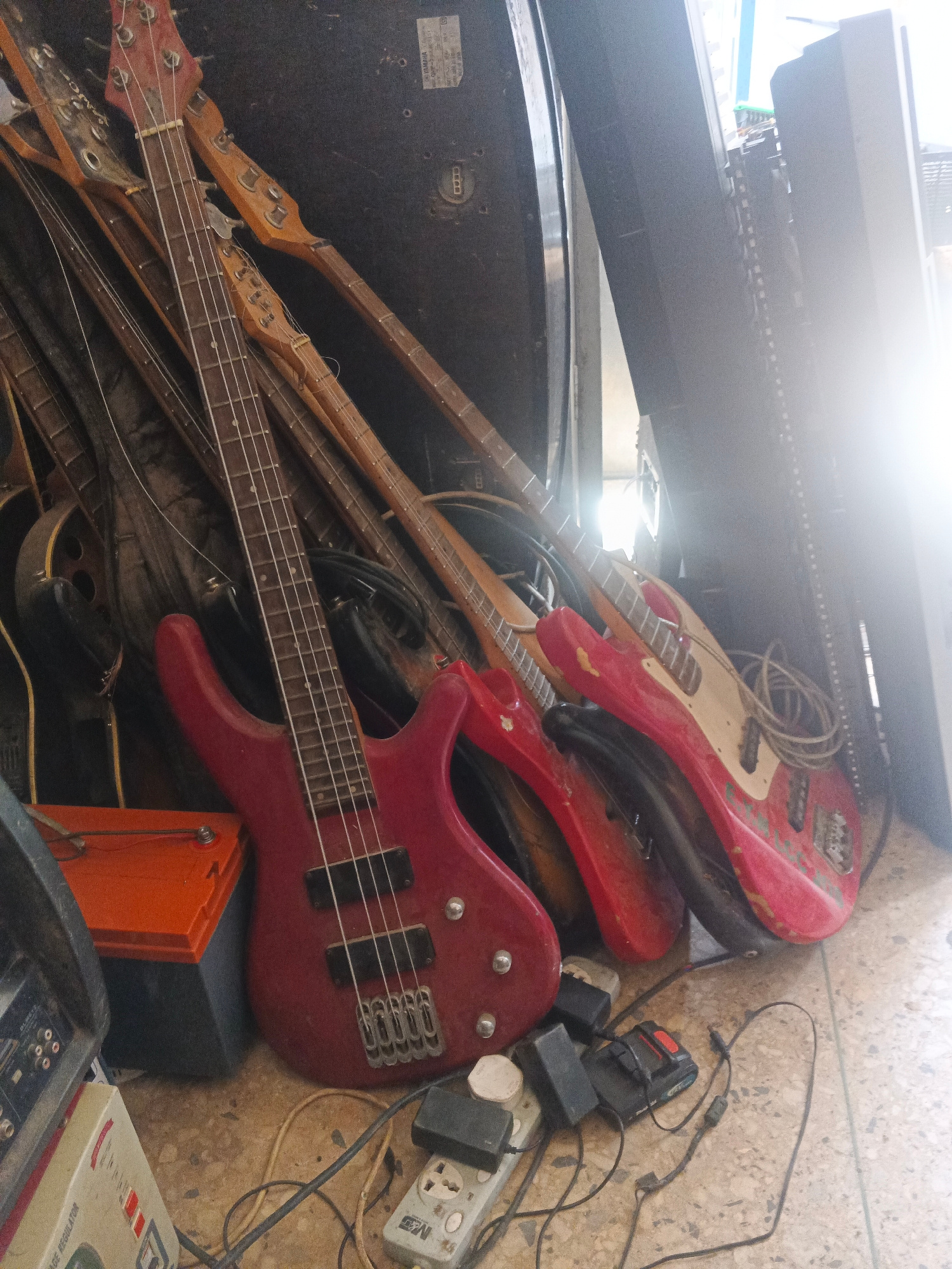 This image depicts a cluttered storage area, likely a workshop or a storage room, filled with musical instruments and electronic equipment. The scene appears to be located in Jos, Nigeria, based on context not visible in the image itself.
In the foreground, a vibrant red bass guitar is prominently displayed, leaning against other instruments. Its fretboard and four strings are clearly visible. To its left is an orange battery or power bank, and further left, a black electronic device with multiple ports. Various cables and electrical adapters are scattered on the floor around these items, suggesting an active or recently used space.
Behind the red bass, several other guitars and basses are stacked, some with reddish-pink bodies and others with natural wood finishes. The instruments are leaning against a large, dark, possibly black, vertical structure that could be a piece of equipment or a wall. A bright light source, possibly sunlight from a window, is strongly illuminating the upper right portion of the image, creating lens flare and washing out details in that area. The floor is tiled with a light-colored, speckled pattern. The overall impression is one of a disorganized but functional space dedicated to music production or repair.