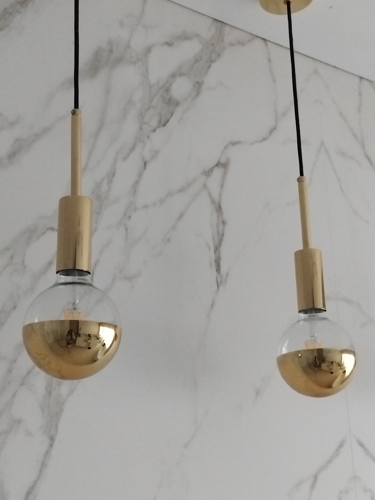 The image shows two gold-dipped globe light bulbs hanging from black cords against a white marble-patterned wall. The bulbs are partially submerged in gold, with the filament visible in the clear upper portion. The wall has a subtle, veined marble pattern. The lighting appears to be natural, possibly from a window, casting reflections on the gold surface of the bulbs. No people or specific locations in Vila Nova de Famalicão, Portugal are visible.