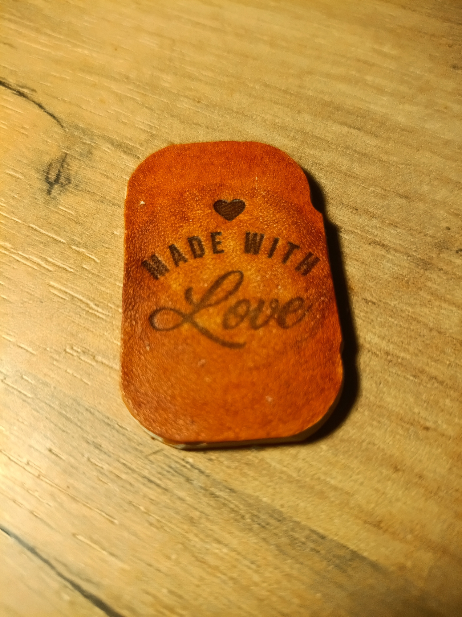 The image shows a single, rectangular, orange leather tag with rounded corners, placed on a light brown wooden surface. The tag is embossed with the text "MADE WITH LOVE" and a small heart above the word "WITH". The wooden surface has a visible grain pattern and appears to be a table or countertop. The lighting casts a shadow from the tag onto the surface. There are no people or other objects visible in the image. The location in the city of Irkutsk, Russia, is not discernible from the image itself.