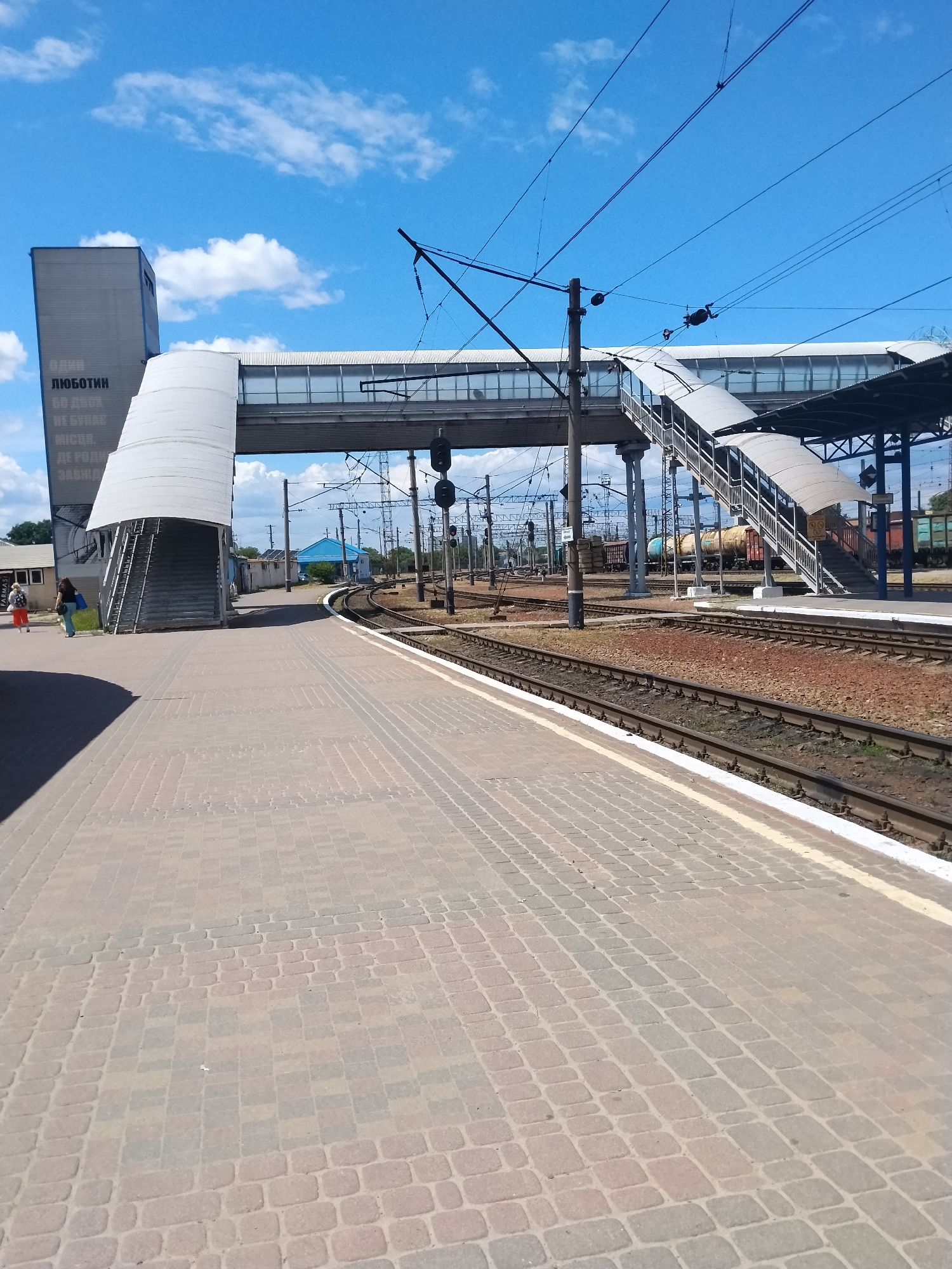 This image captures a train station platform under a partly cloudy blue sky. The platform itself is paved with light brown and grey bricks laid in a cobblestone pattern. To the left, a modern, multi-story building with a grey facade and a white, curved canopy over an outdoor staircase is visible. Text in an unknown language is present on the building. Two individuals, one wearing an orange skirt and the other a blue shirt, are walking on the platform away from the camera. Across the platform, multiple train tracks stretch into the distance, bordered by gravel and ballast. Overhead, a network of power lines and supporting structures for an elevated pedestrian bridge are visible. The bridge, enclosed with glass, spans across the tracks, with visible staircases on either side. In the background, several train cars, including a tanker car, are parked on the tracks, and a small blue building with a pitched roof is situated amongst trees. The scene appears to be a functional, active train station environment.