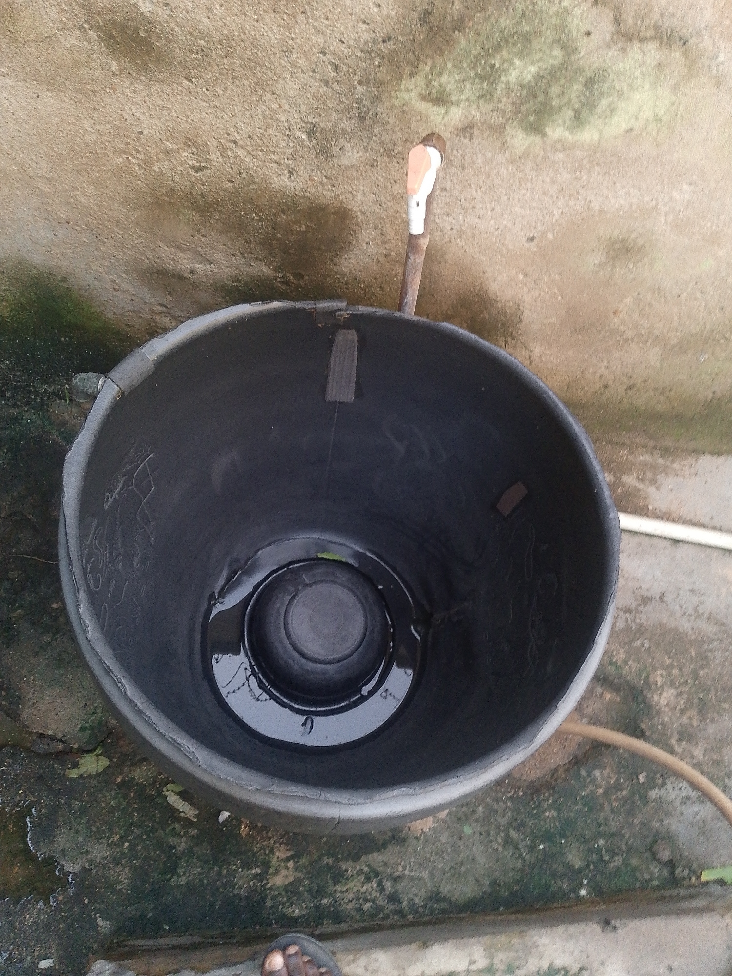 The image shows a black plastic bucket sitting on a concrete surface, possibly outdoors. Inside the bucket, there is a small amount of water, with a darker circular object submerged in it. The bucket's rim is worn and shows signs of use. A thin pipe, possibly a faucet or part of a water system, is visible extending from the wall behind the bucket, with a white and orange handle. A brown hose is also seen curving away from the bucket towards the right side of the frame. The concrete surface around the bucket appears damp and has some patches of green moss or algae, suggesting a humid environment or recent rain. In the bottom right corner, a person's foot wearing a sandal is visible, indicating that a person is present, but only their foot is in the frame. The overall lighting suggests it could be daytime.