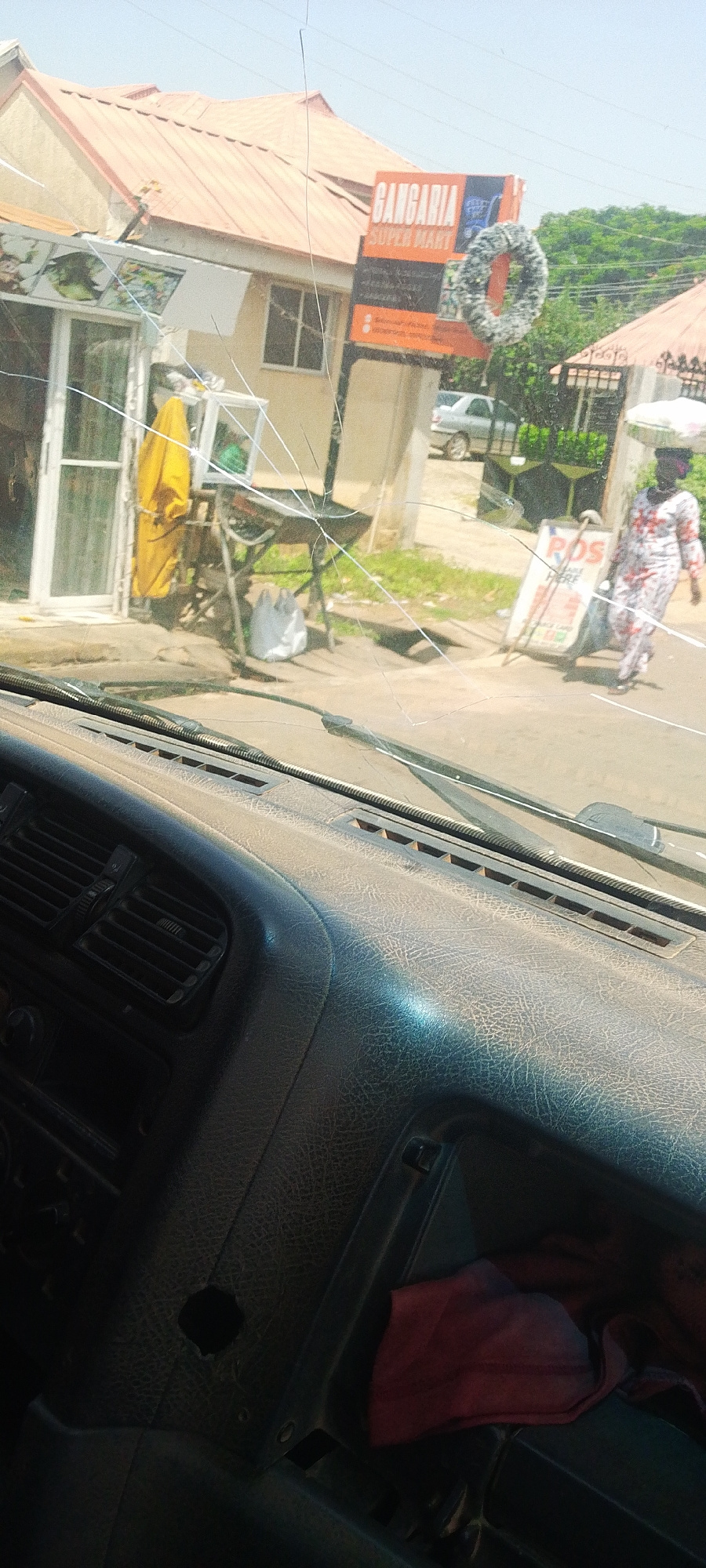 The image shows a view from inside a vehicle, looking out through a cracked windshield at a street scene in Jos, Nigeria. The foreground is dominated by the dashboard of the vehicle, which appears worn and dusty, with visible vents and a dark-colored material.
Outside, the scene suggests a typical street in a warm climate. A building with a red-tiled roof and cream-colored walls stands to the left, housing a shop with a glass storefront. A bright yellow garment hangs on display inside or near the shop entrance. To the right of the shop, a metal stand holds a mirror and other items. Further down the street, a woman walks with a large basket balanced on her head, wearing a patterned dress and headscarf. A white sign with red lettering that appears to read "POS HERE" is visible near her.
Above the shop, a large sign advertises "GANGARIA SUPER MART." The sign is orange and black, with the words "GANGARIA" prominently displayed in white lettering, followed by "SUPER MART" in smaller white letters. A decorative wreath-like object, possibly made of faux fur or tinsel, hangs from the sign.
The sky is bright and clear, indicating a sunny day, likely mid-morning or afternoon. The presence of lush green trees in the background suggests an area with vegetation. The overall impression is of a bustling commercial street in a Nigerian town. The cracked windshield adds a sense of ruggedness or perhaps signifies a recent event, though no specific incident is depicted.
