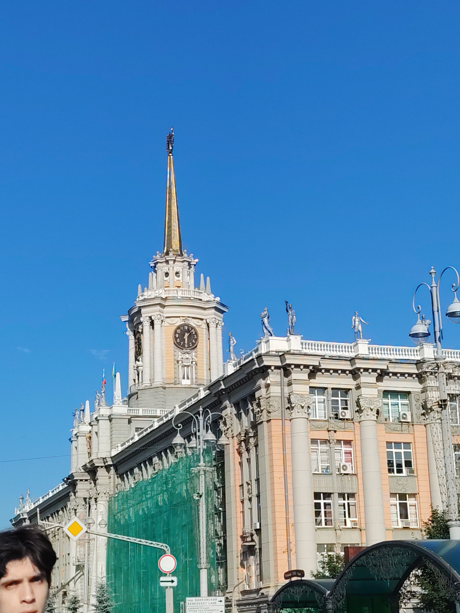 This image captures an architectural marvel against a clear blue sky, hinting at a bright, sunny day. The scene is dominated by a grand, multi-story building with classical influences, featuring ornate columns, intricate detailing, and a prominent clock tower topped with a golden spire. Several statues of figures are strategically placed along the building's roofline, adding to its majestic appearance.
The building's facade displays a mix of architectural styles. The lower sections are adorned with peach-colored elements and large columns, while the upper sections are lighter in color, with white detailing and balustrades. A large section of the building is covered by green netting, suggesting ongoing renovation or construction work.
In the foreground, the top of a person's head and face are visible, looking upwards, suggesting they are observing the architecture. The person appears to be a young man with dark hair.
In the background, a street light with a classic design is visible to the right, and road signs can be seen, including one with Cyrillic text. One sign in the lower right corner has the text "пер. БАНКОВСКИЙ" and "ГОРОДА ЕКАТЕРИНБУРГА" which indicates the location as Yekaterinburg, Russia, specifically Bankovsky Lane. The overall impression is one of civic pride and historical grandeur, possibly in a city square or a significant urban area.