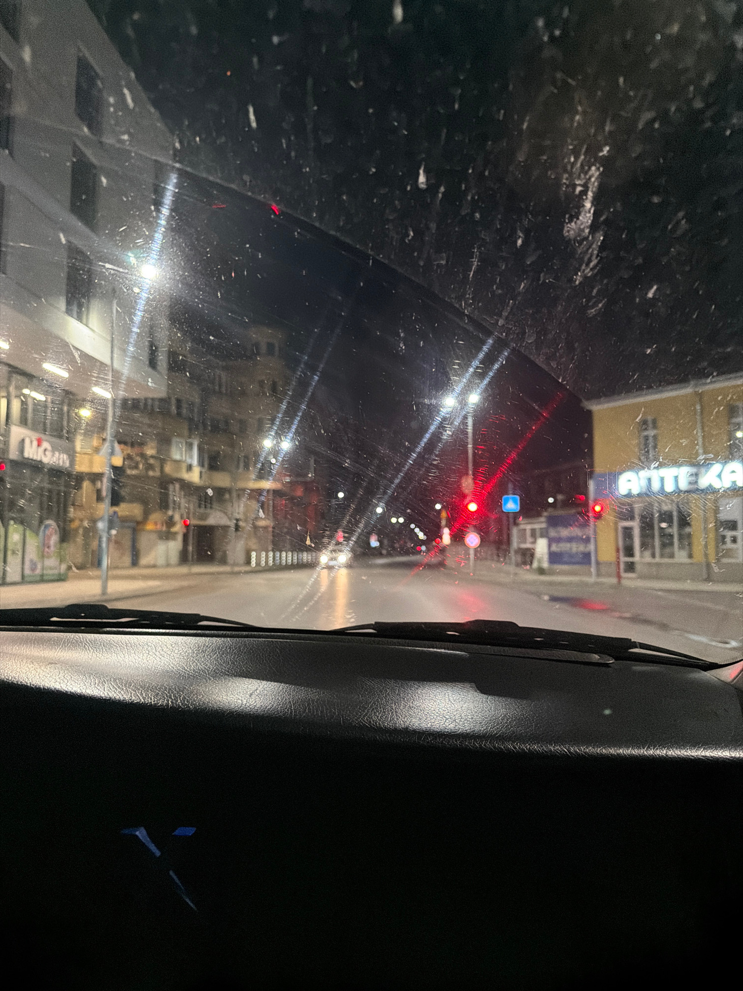 The image shows a view from inside a car at night, looking through a wet windshield. The car appears to be driving on a city street in Plovdiv, Bulgaria, indicated by the signage. The street is lined with multi-story buildings on both sides, many of which are illuminated. There are streetlights and traffic lights visible, with several red lights suggesting the car is stopped or approaching a red light. One building on the right has a prominent sign that reads "ANTEKA." The windshield is covered in raindrops and streaks, distorting the view of the street and buildings. The foreground of the image shows the dark dashboard of the car. The overall scene depicts a nighttime drive through a European city, likely in the rain.
