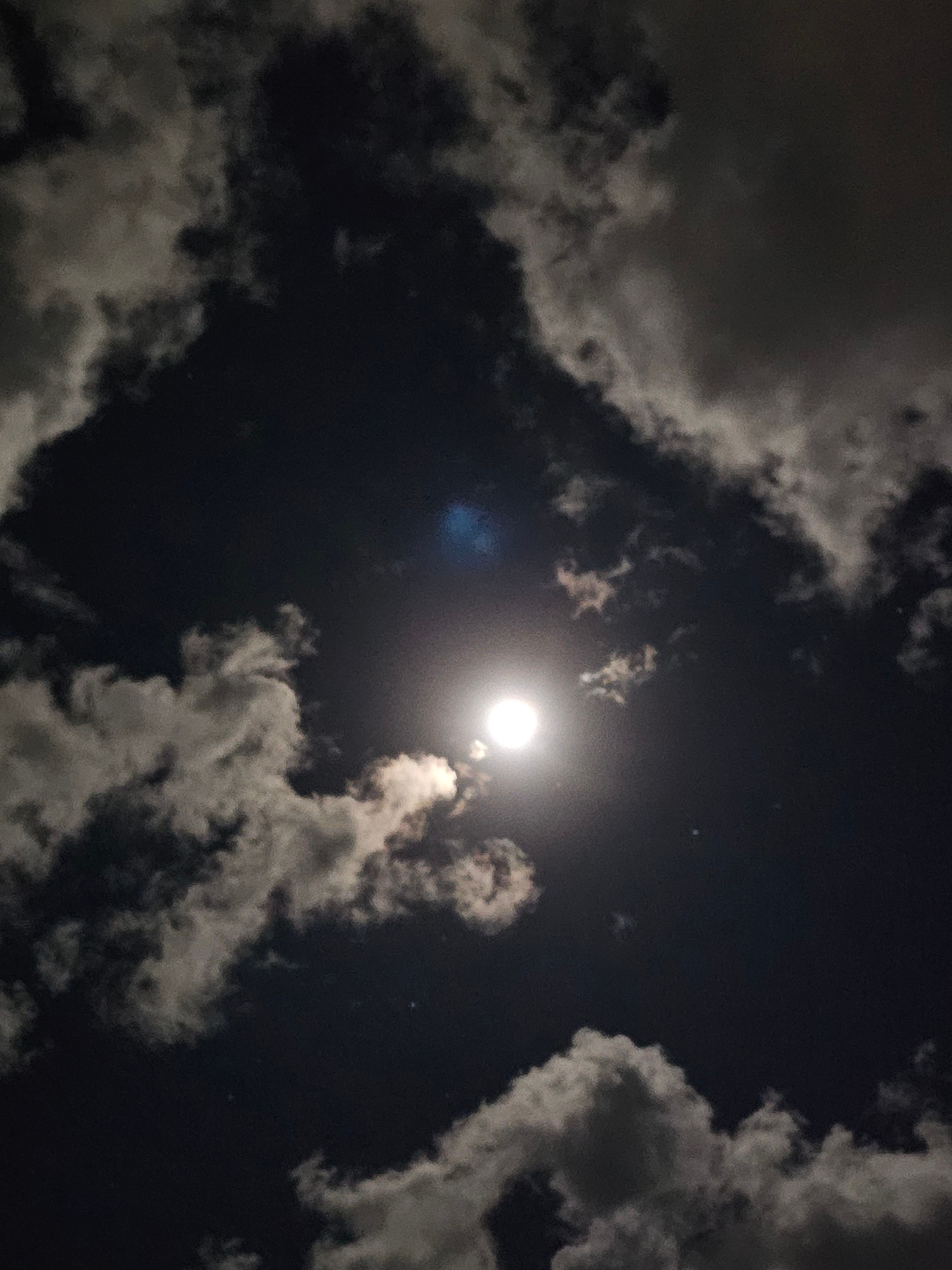 The image shows a night sky filled with clouds and the moon. The moon is bright and centrally located, casting a soft glow through the clouds. Some clouds appear dark and dense, while others are illuminated by the moonlight, creating a dramatic and somewhat hazy effect. The sky is dark, indicating nighttime. There are no people or man-made objects visible, suggesting a natural outdoor setting. The overall mood is serene and mystical, evoked by the interplay of light and shadow in the night sky. The location context of Denpasar, Indonesia, places this scene in a tropical environment, though no specific geographical features are discernible in the image itself. The weather appears to be partly cloudy. There is no visible text in the image.