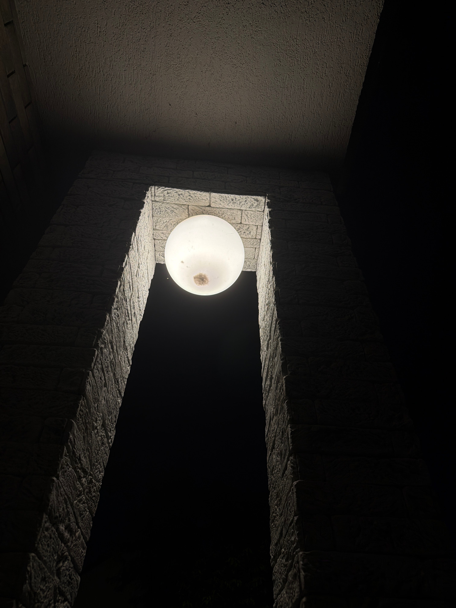 A low-angle, close-up shot captures a spherical, illuminated light fixture hanging within a stone archway at night. The light fixture is off-white and appears to have a small, discolored area in its center. The archway is constructed from light-colored stone blocks with a textured surface, resembling a rough-hewn brick pattern. The stone elements are illuminated by the light fixture, creating stark shadows and highlighting the texture. The background is predominantly black, indicating a dark outdoor environment, possibly with faint foliage visible at the bottom edge. The ceiling above the archway is also visible, appearing textured and painted in a neutral tone. The image is devoid of any human presence or activity, focusing solely on the architectural feature and its lighting. The overall impression is one of an outdoor architectural detail, possibly an entrance or decorative element, illuminated for nighttime visibility.