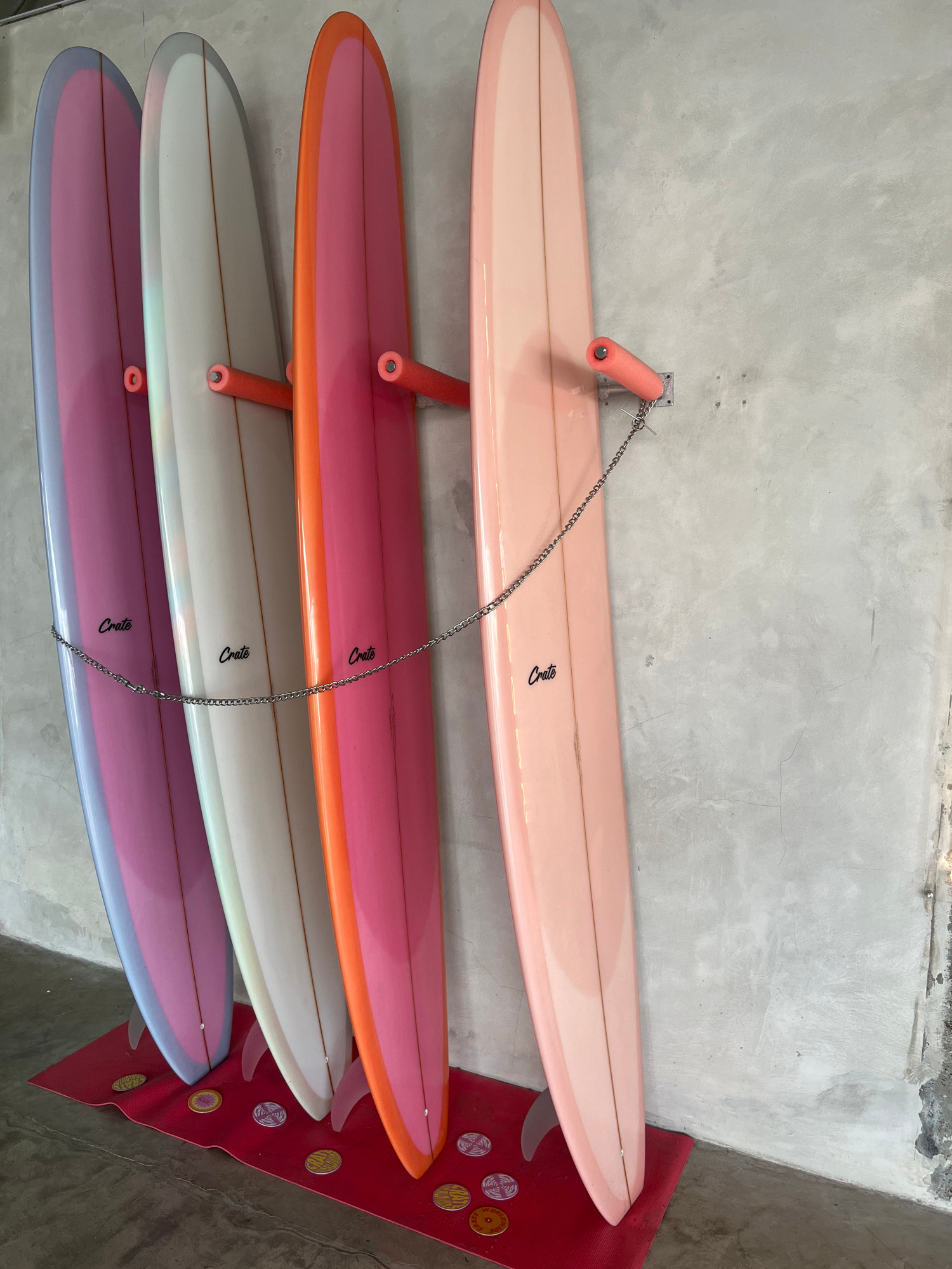 Four longboard surfboards are displayed vertically, leaning against a textured, grey concrete wall. The surfboards are arranged from left to right in the following colors: blue-grey and pink gradient, white with grey trim, orange and pink gradient, and a pale peach. Each surfboard has the word "Crate" written in black cursive script. A silver chain is looped around the surfboards and attached to orange foam-covered wall hooks. The surfboards rest on a red mat, which is on a grey concrete floor. Small, circular decorative discs are scattered on the mat. The scene is indoors. The location is specified as Mengwi, Indonesia.