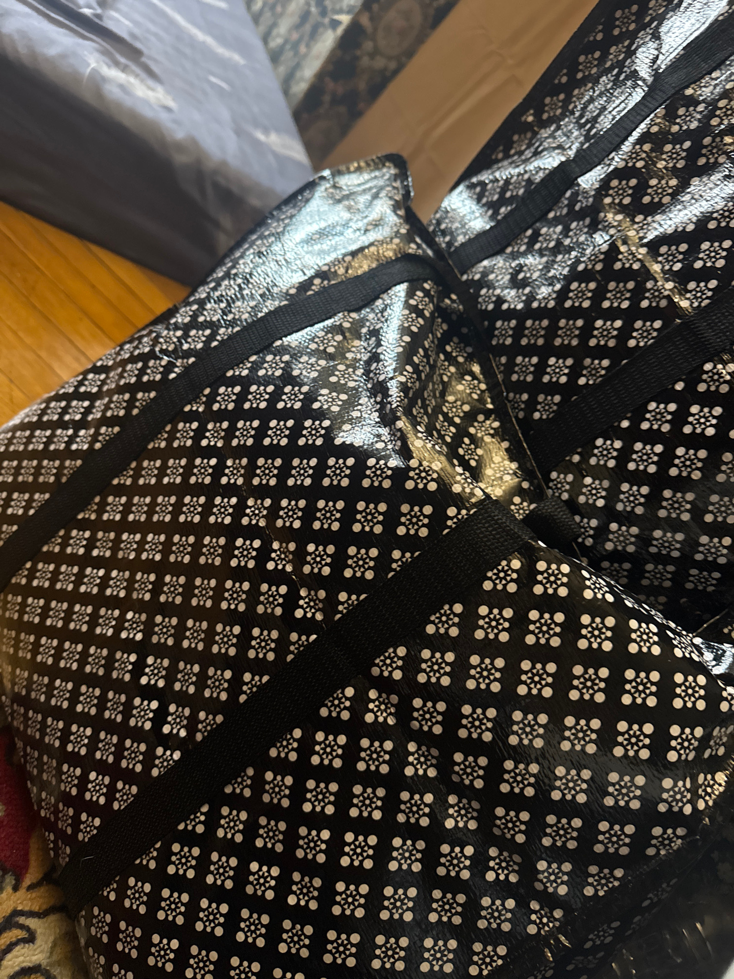 The image shows two large, black reusable shopping bags with a white geometric pattern of small circles and triangles arranged in a flower-like shape. The bags are made of a shiny, possibly laminated material. One bag is placed on top of the other, and both have black fabric straps. They are positioned on a wooden floor next to a dark grey, possibly upholstered object, and a patterned rug. There are no people or specific Moscow landmarks visible.