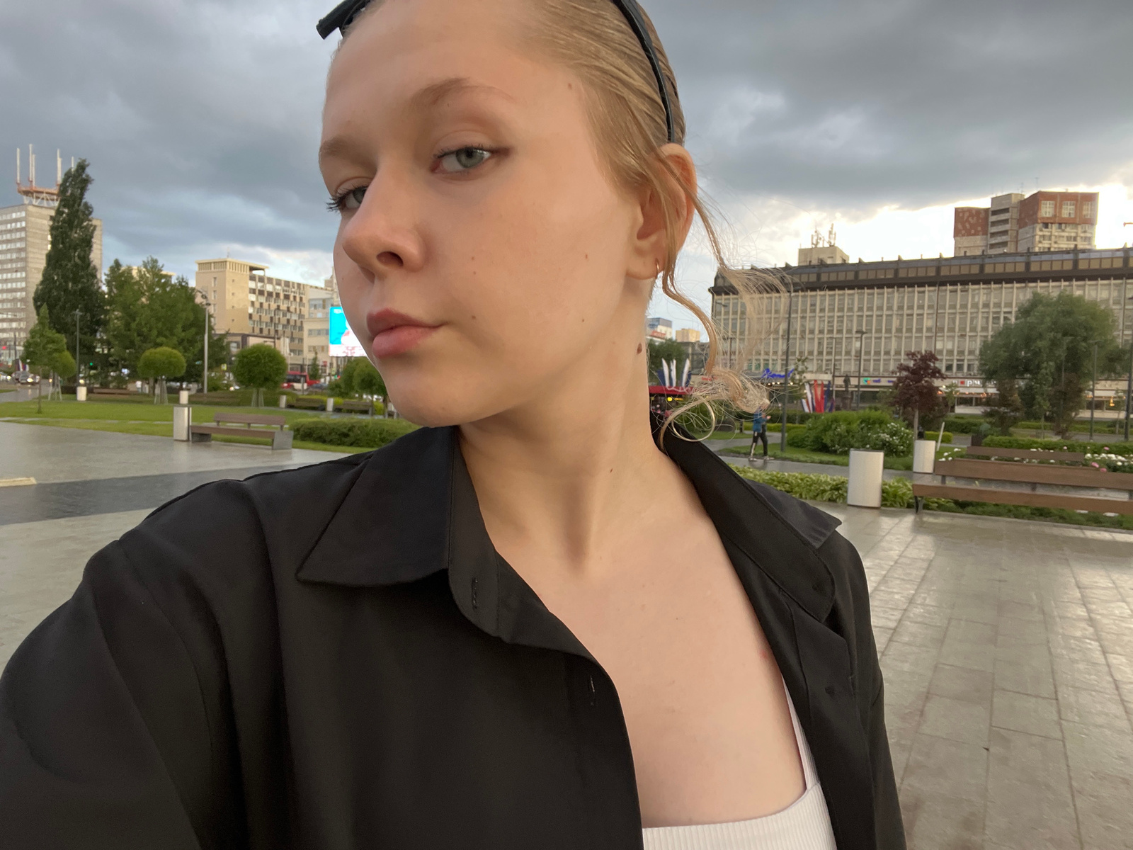 A young Caucasian woman with blonde hair pulled back by a black headband and wearing a black open button-up shirt over a white tank top is looking towards the camera. Her face is turned slightly to the left. The background shows an urban environment with modern buildings and trees under a cloudy sky. A paved walkway and park benches are visible in the foreground. The image appears to be taken in the city of Perm, Russia, possibly near a central square or park area.
