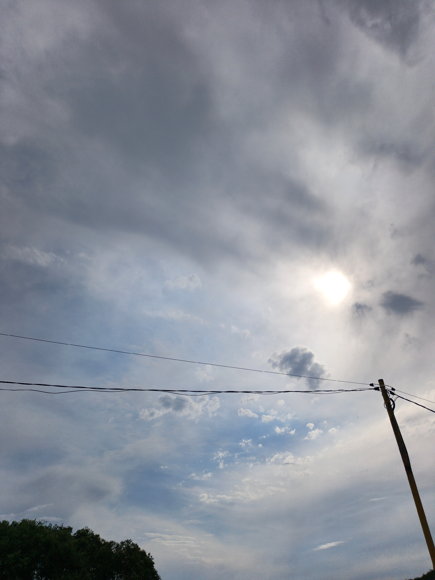 The image depicts a cloudy sky with the sun peeking through, creating a bright halo effect. Two power lines stretch horizontally across the frame, supported by a wooden utility pole on the right side. The pole appears weathered and is angled slightly towards the viewer. In the lower left corner, the dark green foliage of trees is visible, suggesting a natural landscape. The overall lighting and the presence of clouds indicate it might be late afternoon or early evening, with a mix of sunlight and overcast conditions. No people or text are visible in the image. The scene conveys a sense of natural elements and man-made infrastructure coexisting.