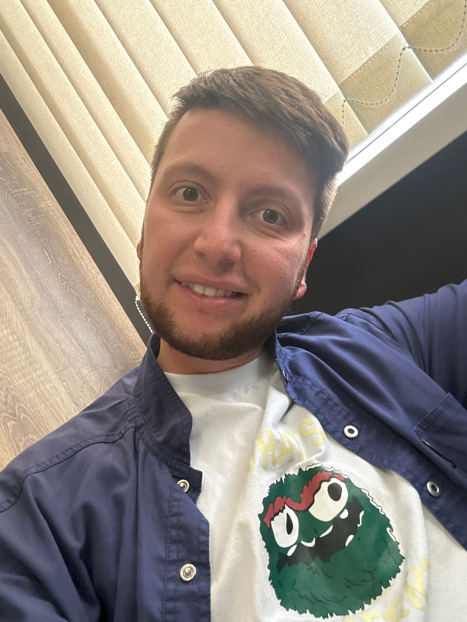The image is a close-up, high-angle selfie of a young man with short brown hair and a beard, smiling. He is wearing a dark blue collared shirt unbuttoned over a white t-shirt with a cartoonish green monster graphic. The background appears to be an office environment with vertical blinds and a wooden-textured surface. There is no indication of the location in Moscow, Russia.