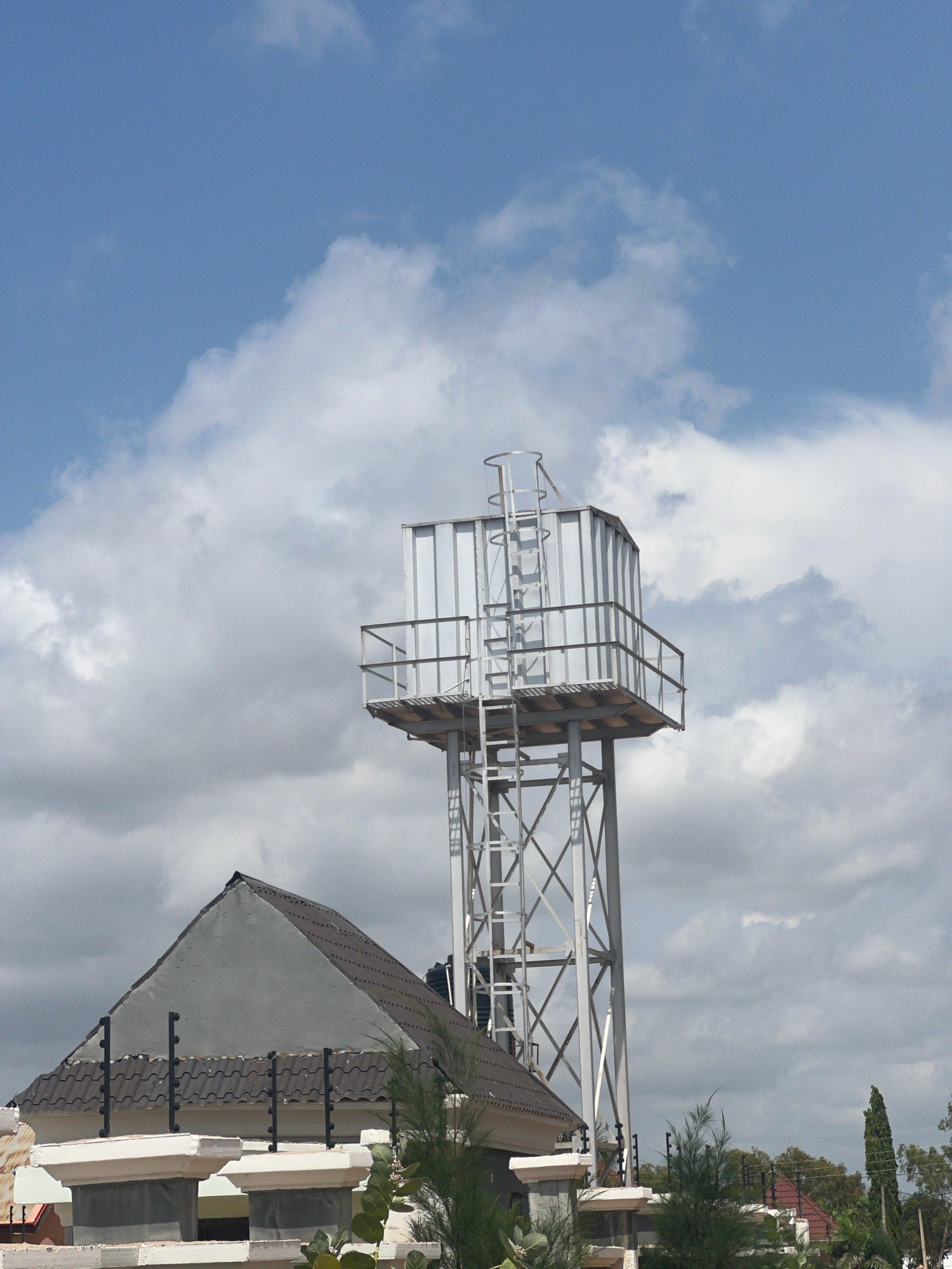 This media file depicts an outdoor scene featuring a large, empty billboard structure against a partly cloudy sky. The billboard is constructed from metal beams and appears to be in a state of disrepair, with some sections missing or damaged. Below and to the right of the billboard, part of a building with a tiled roof is visible. The building has a distinctive architectural style with a pointed roof section and decorative elements. Lush green trees and foliage surround the building, suggesting a tropical or subtropical environment.
The time of day is difficult to determine precisely, but the lighting suggests it could be late morning or early afternoon. The weather appears to be mixed, with patches of blue sky interspersed with cumulus clouds, indicating fair weather with some cloud cover.
While no people are visible in the frame, the presence of the billboard and building implies a human-made environment, likely a roadside or a commercial area in Howayi, Nigeria, as indicated by the location context. The scene is static, with no discernible activity taking place. The overall mood is neutral, with the focus on the architectural elements and the natural surroundings. There is no visible text in the image.