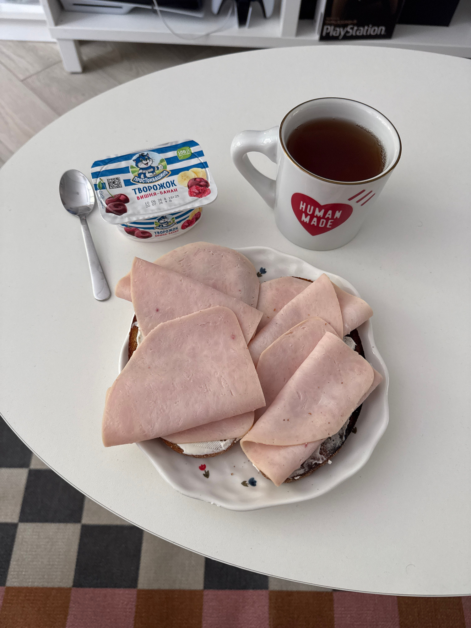 A top-down view shows a white round coffee table with breakfast items. On the left, a small, round container of yogurt with a blue and white striped lid is placed next to a silver spoon. The yogurt container has Cyrillic text and images of cherries and bananas. To the right of the yogurt is a white mug with a red heart design and the words "HUMAN MADE" in red lettering. The mug contains a dark liquid, likely tea. In the center of the table is a white plate with a small floral design. On the plate, there is a slice of dark bread topped with a white spread and several thin slices of pink ham. The background shows a portion of a white shelf unit and a black PlayStation game case. The table is situated on a checkered rug with black, grey, brown, and pink squares. The scene suggests a casual breakfast setting.
