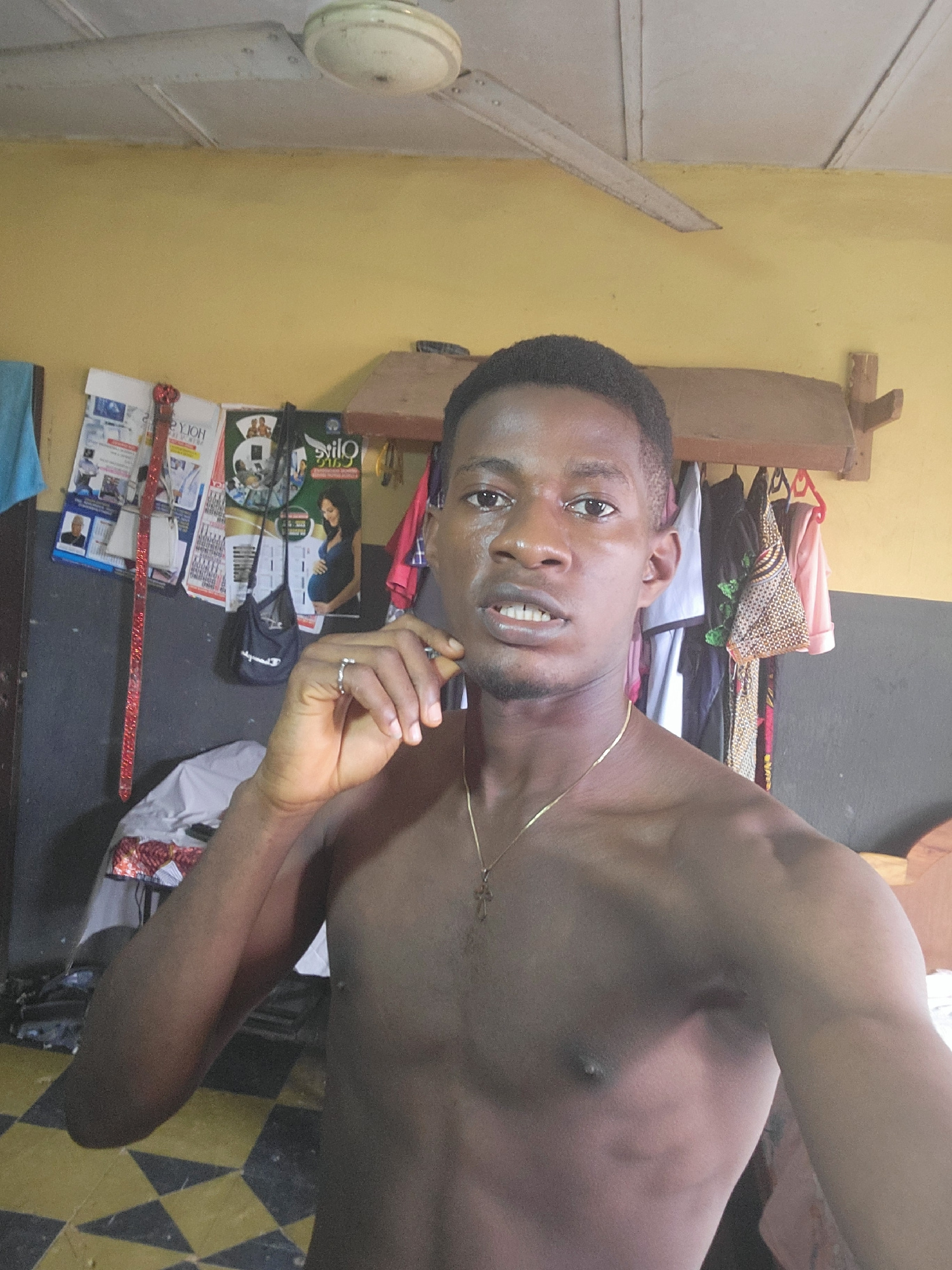 A young man is pictured from the chest up, looking directly at the camera. He is shirtless and wearing a thin gold chain with a cross pendant. His right hand is raised to his face, with his ring finger touching his chin. He has dark hair and appears to be in a room with yellow walls and a tiled floor. There are posters and clothing visible in the background. A ceiling fan is mounted on the ceiling. The image appears to be a selfie.