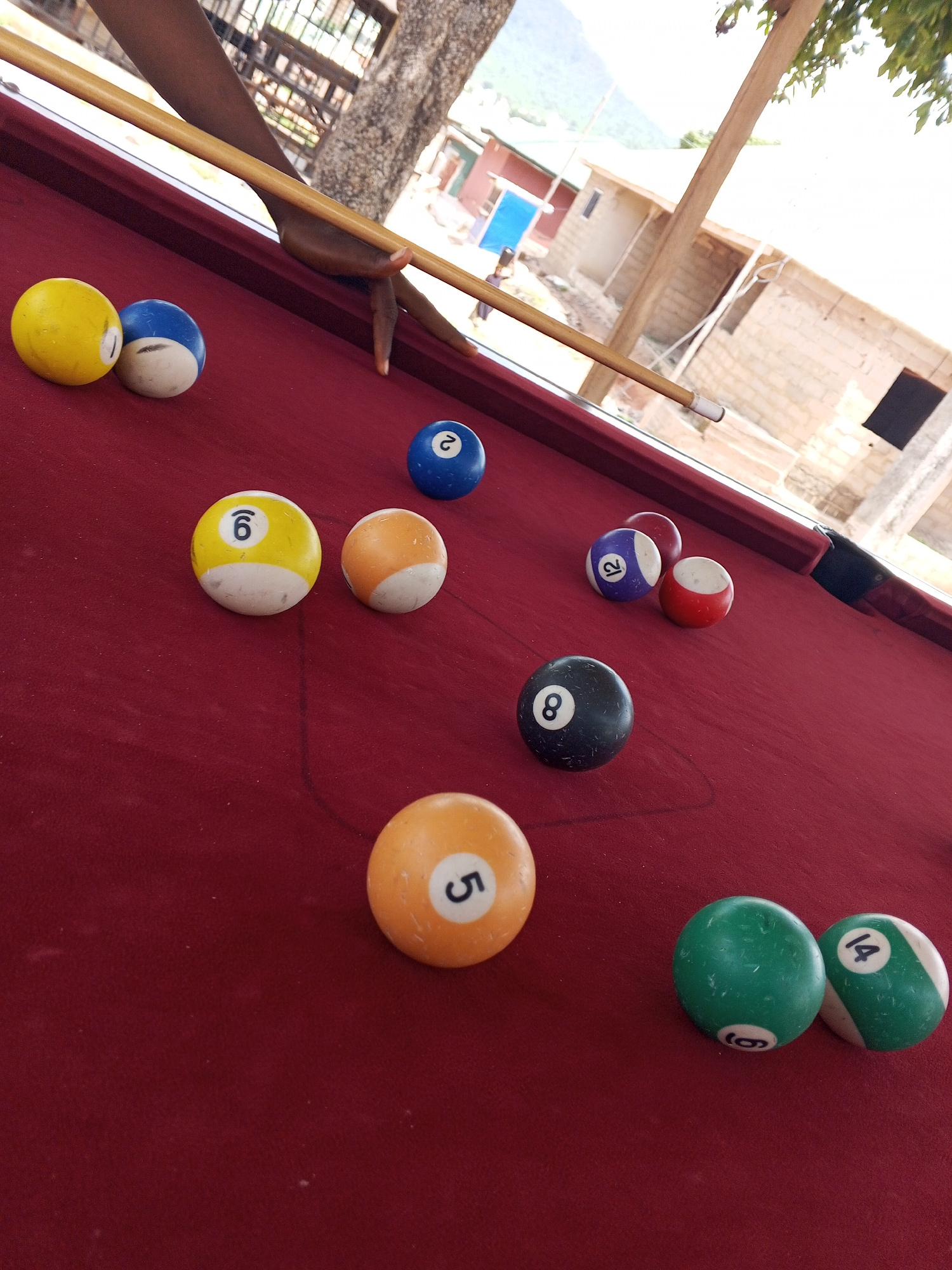 A close-up, low-angle shot captures a pool table on a red felt surface outdoors, likely in Lokoja, Nigeria, given the background scenery of buildings and mountainous terrain bathed in bright sunlight. A person's arm, dark-skinned, is visible holding a pool cue, poised to strike a billiard ball. Several pool balls are scattered on the table: a yellow ball with the number 1, a blue and white ball with a partial number, a blue ball with the number 2, a yellow and white ball with the number 6, an orange and white ball, a purple ball with the number 12, a red and white ball, a black ball with the number 8, an orange ball with the number 5, a green ball with the number 6, and a green and white ball with the number 14. The background reveals a rustic environment with several low-rise buildings constructed from concrete blocks, some with corrugated iron roofs, and sparse vegetation. The overall impression is of a casual game of pool being played in a relatively undeveloped outdoor setting.