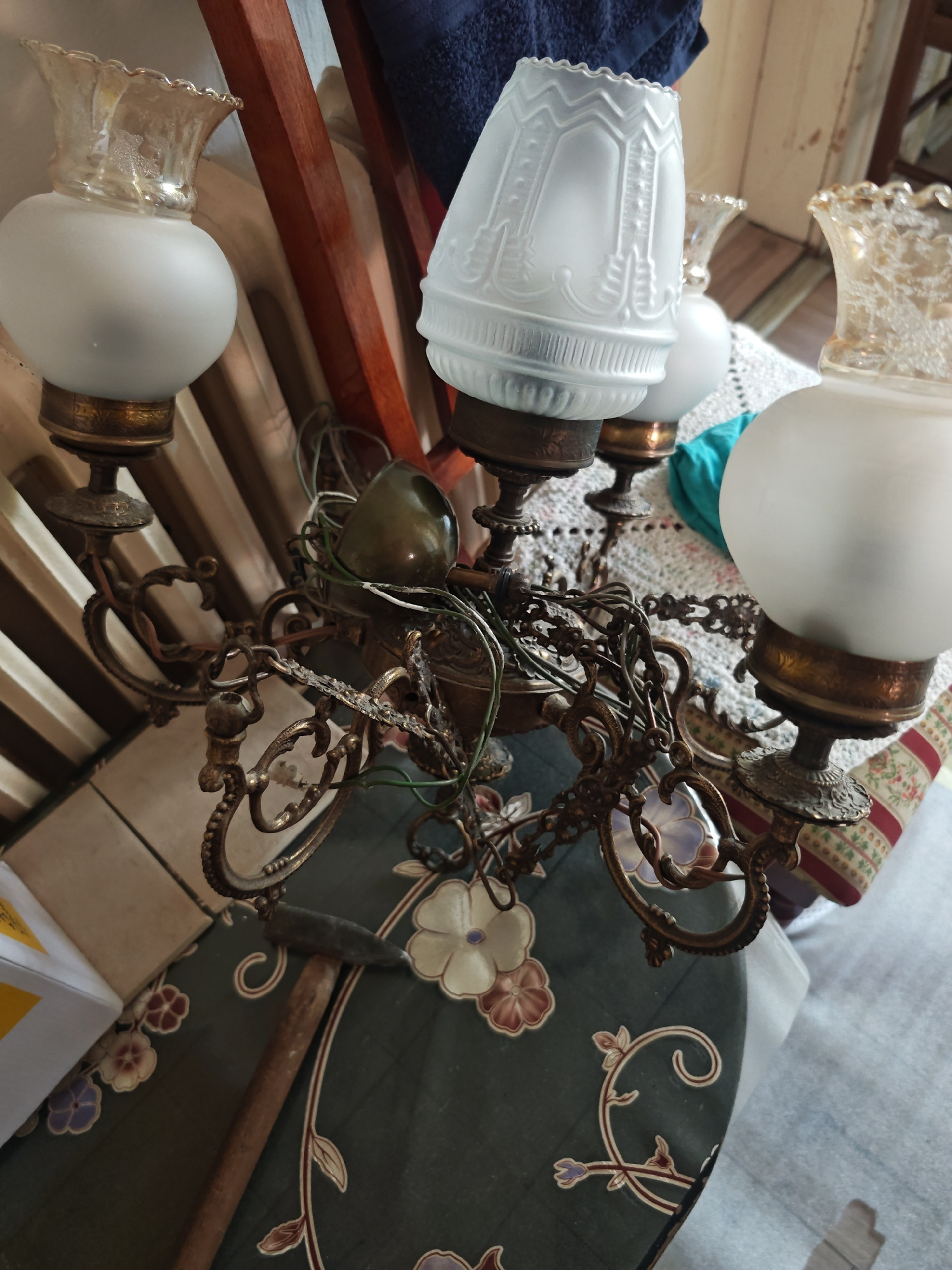 A close-up, slightly angled shot shows a vintage brass chandelier with four glass shades and some exposed wiring. The chandelier has intricate metalwork, including chains and decorative arms. One of the shades is frosted and conical, while the other three are globe-shaped with ornate glass tops. A green, tear-drop-shaped object is visible among the wiring. The chandelier is positioned above a dark green ironing board with a floral pattern. To the left of the chandelier, a small hammer with a wooden handle rests on the ironing board. In the background, a wooden structure, a dark blue textile, and a white crocheted blanket are partially visible. The lighting is subdued, suggesting an indoor environment. The location is identified as Unknown, Unknown.