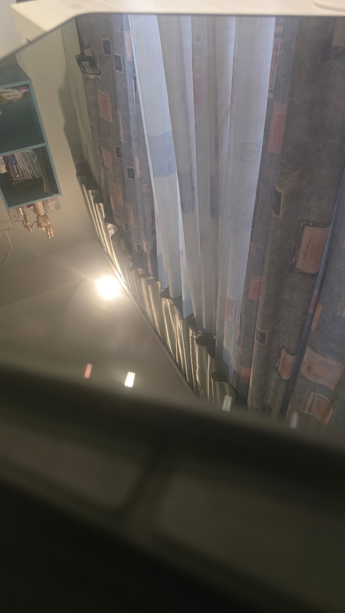 The image shows a reflection on a dark, possibly glass surface, with a bright light source creating a glare. In the reflection, there are patterned curtains, a shelf with books, and a light fixture. The scene appears to be indoors, possibly in a room with a window. The city of Ukhta, Russia is not mentioned or visible in the image.