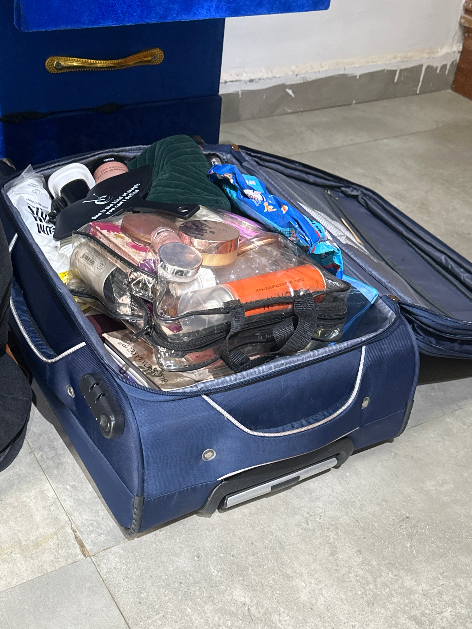 In Abuja, Nigeria, a navy blue suitcase lies open on a grey tiled floor, revealing its contents. The suitcase is packed with various personal items, primarily cosmetics and toiletries, housed in clear plastic bags and scattered amongst other objects. Notably visible are several bottles of makeup, skincare products, and what appears to be a makeup brush. The suitcase itself has a telescoping handle and what looks like a built-in lock.
To the left of the suitcase, a part of a dark blue sofa with a decorative gold handle is visible. The surrounding environment suggests an indoor setting, possibly a bedroom or a living area, with unfinished or textured walls in the background. The lighting appears to be artificial, casting some shadows. Text visible on some of the items includes brand names like "BLOSSOM MAKEUP," "MAREUE," and "LOCKING." Other text includes phrases like "TOTALLY SMUDGE & TRANSFER PROOF," "New In," "you can't define," and "She is the kind of magic," indicating a focus on beauty products. The scene captures the preparation for or unpacking of travel essentials, with the contents suggesting a personal journey.