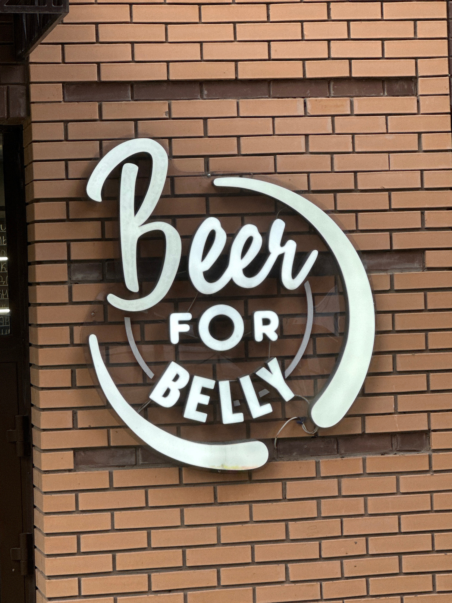 A close-up shot shows a lit sign with white, stylized lettering on a brick wall. The sign reads "BEER FOR BELLY" in a circular design. The brick wall is made of light brown bricks laid in a stretcher bond pattern. The sign itself is white and appears to be illuminated from within. The lighting suggests it might be daytime or that the sign is lit even during the day. The bricks are varied in color, with some being a lighter tan and others a darker reddish-brown. The mortar between the bricks is a darker gray. The sign has a curved shape that follows the letters. A black metal shelf or structural element is partially visible at the bottom left of the image, and a dark band runs along the bottom edge, potentially indicating the top of a window or doorway. The image is taken from a slightly upward angle. The overall setting appears to be the exterior of a building, likely a business. There are no people visible. The location is identified as Irkutsk, Russia.