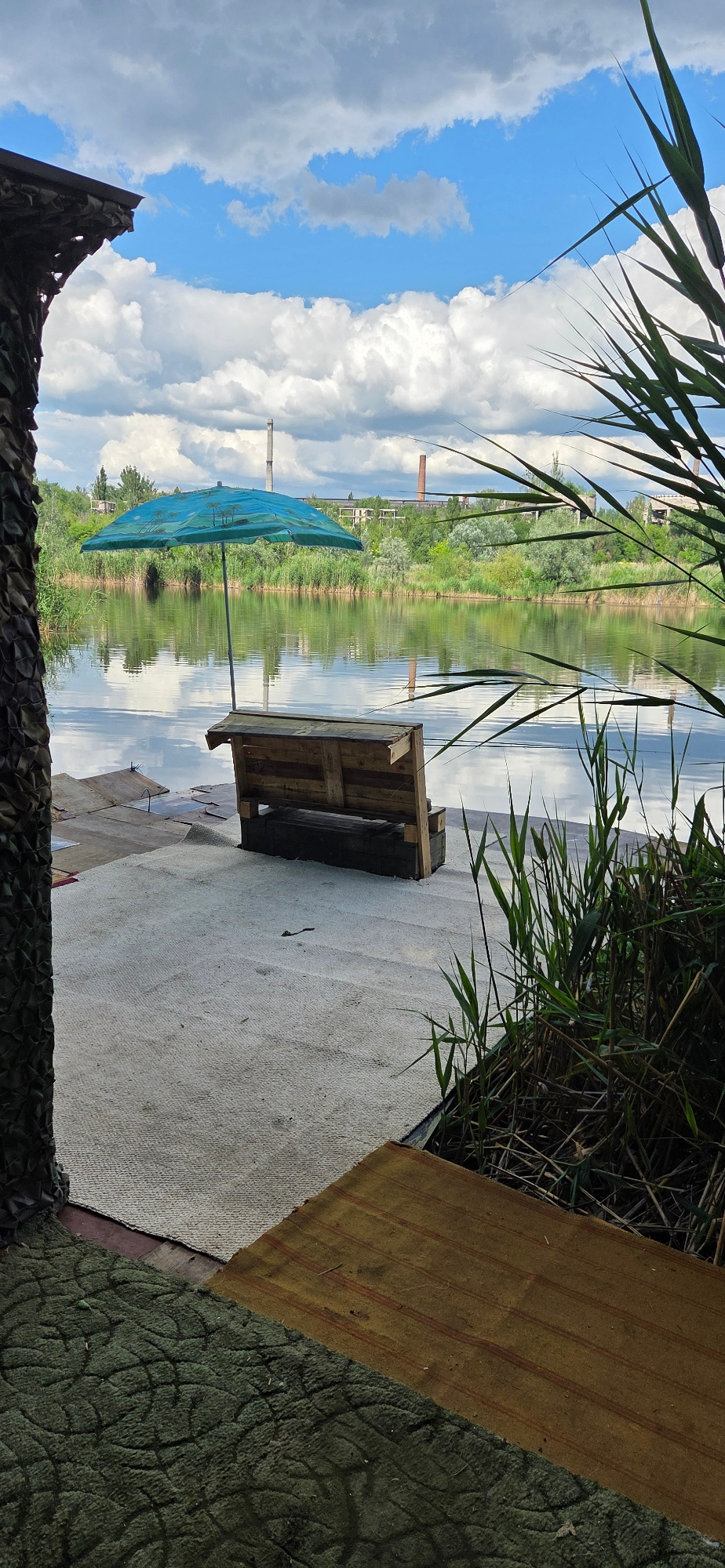 This is a landscape-oriented, slightly tilted, outdoor photograph featuring a serene waterside scene. The foreground is dominated by a textured, light-colored surface that appears to be canvas or a similar material, possibly part of a makeshift seating area. To the left, a dark, patterned camouflage netting peeks into the frame, and to the right, there is a diagonal strip of textured brown material with red stripes, possibly wood or cardboard, adjacent to a dark green textured surface.
Behind this foreground, a wooden pallet structure is visible, serving as a base for something unseen. Further back, a blue umbrella with a tropical pattern is propped open, providing shade. This umbrella stands near the edge of a calm body of water.
The water reflects the sky and the lush green vegetation lining the opposite bank. The far bank is characterized by dense greenery, with scattered trees and evidence of some industrial or abandoned structures, including brick chimneys. The sky above is a vibrant blue, dotted with fluffy white clouds, suggesting a bright, possibly sunny day. Tall, thin reeds or grass blades frame the top of the image, casting shadows and adding to the natural ambiance.
The image appears to be taken during daylight, likely in the late morning or early afternoon, given the brightness of the sky and the clear reflections. The overall mood is peaceful and perhaps a bit rustic, hinting at a quiet spot by the water, potentially for relaxation or observation. There is no visible text in the image.