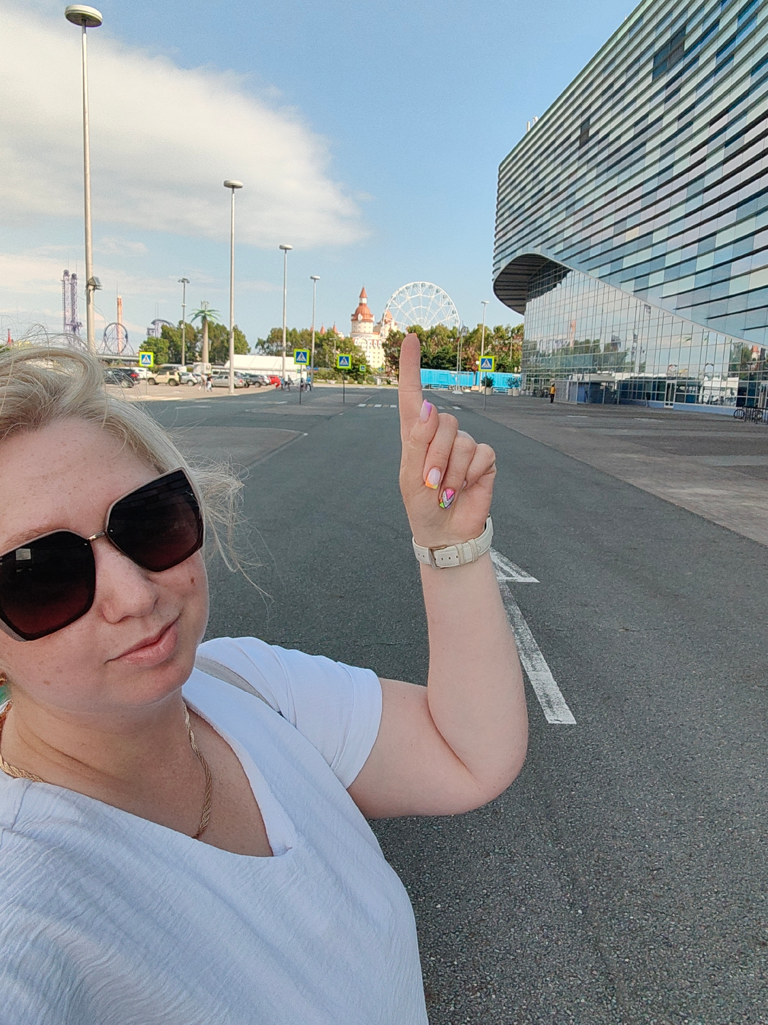 A woman is taking a selfie outdoors, standing on an asphalt surface with white road markings. She is wearing a white t-shirt, large dark sunglasses, and a gold chain necklace. Her left hand is raised, with her index finger pointing upwards. Her nails are painted with a colorful, abstract design. A white watch with a cream-colored band is visible on her left wrist.
In the background, a modern, multi-story building with a facade of horizontal, reflective panels dominates the right side of the frame. To the left of the building, a Ferris wheel is visible, along with the spires of what appears to be a themed park or resort, possibly with roller coasters. Palm trees and other greenery are scattered throughout the scene. Several streetlights and pedestrian crossing signs are also present. A few cars are parked in the distance. The sky is blue with scattered clouds, suggesting a daytime scene with pleasant weather. The overall impression is a vacation or tourist setting, likely at a resort or entertainment complex. The woman's expression is a slight smile.