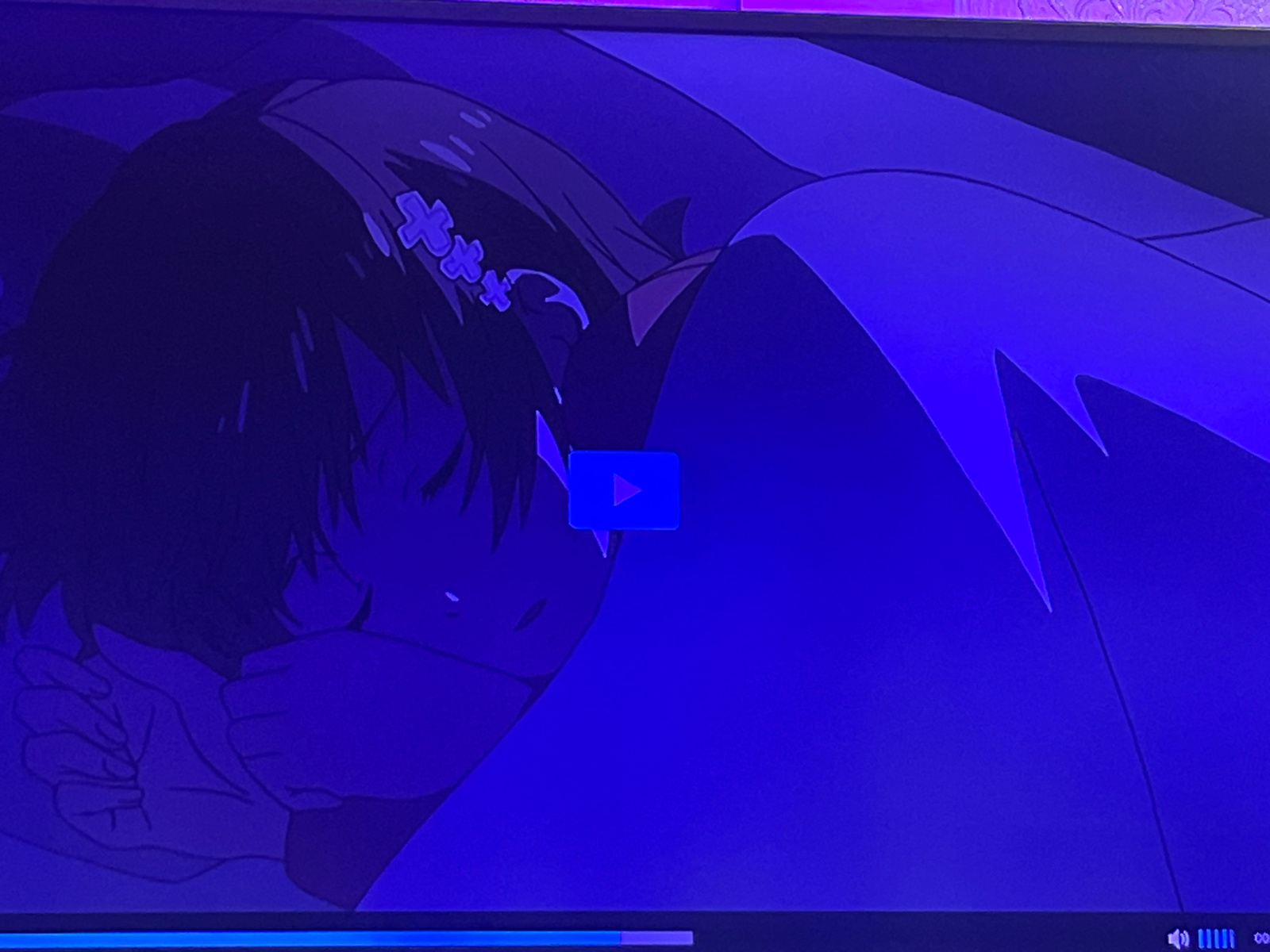 The image is a still from a video player interface displaying an animated scene. The scene depicts a young female character with dark hair, adorned with two purple cross-shaped hair clips, sleeping or resting, with her hands covering her face. The character is under a blanket or sheet, with only her head and upper body visible. The overall color palette of the scene is dominated by deep blues and purples, suggesting a nighttime or dimly lit environment. A blue play button icon is superimposed on the image, indicating that the video is paused or ready to be played. A progress bar is visible at the bottom of the screen, with a partially filled indicator and a volume icon on the right. The image does not contain any identifiable landmarks or elements that would place it specifically in Shahtinsk, Kazakhstan.