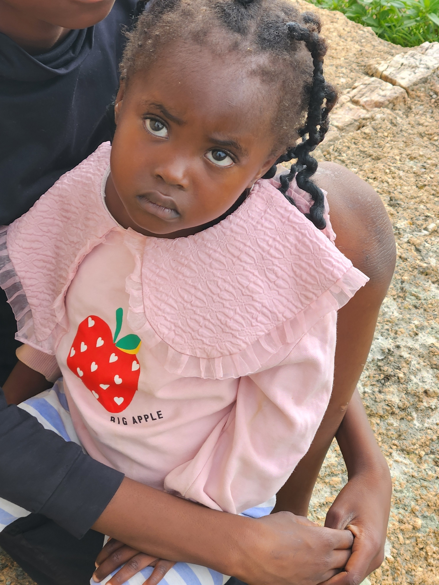 A close-up, eye-level shot features a young Black girl with braided hair, wearing a pink ruffled shirt with a strawberry graphic and the words "BIG APPLE" printed below it, and striped pants. She is being held by an adult, whose dark clothing is partially visible. They are seated outdoors on a rocky surface, with green foliage in the background. The overall scene suggests a casual, possibly familial moment in an outdoor setting. The photographer has likely captured this image within the city of Jos, Nigeria, a location known for its rocky terrain and outdoor spaces.