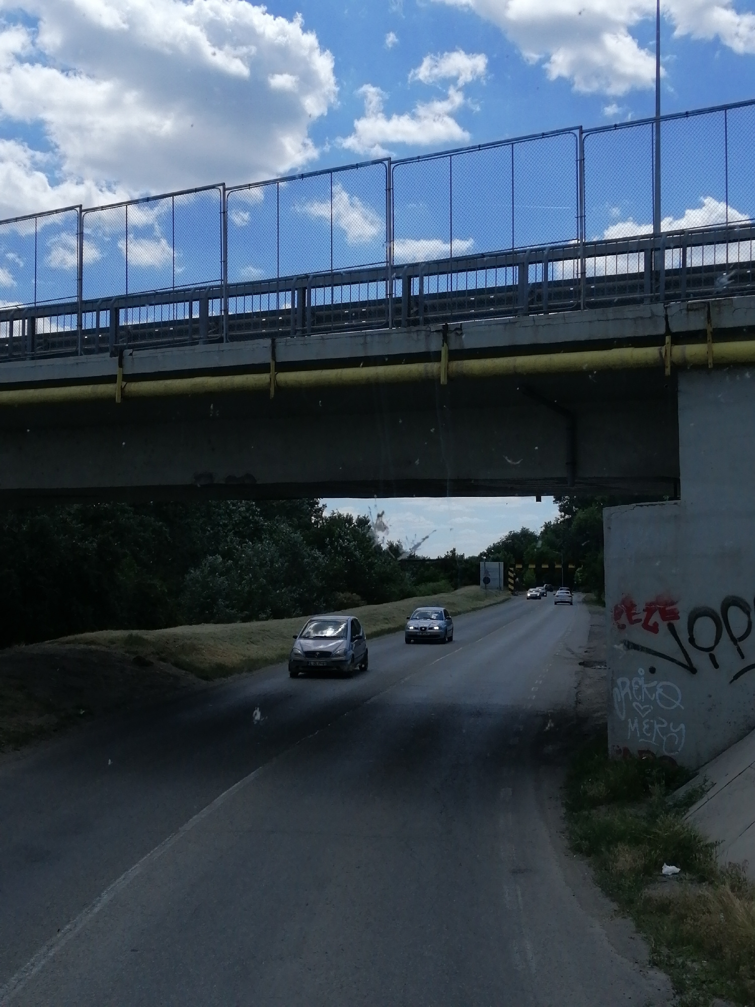 The image captures a view from underneath a bridge, looking out onto a road. In the foreground, a concrete pillar of the bridge is visible on the right, covered in red and white graffiti. The road curves gently to the left and extends into the distance, where trees line the sides. A single silver car is traveling away from the camera on the left side of the road. Further down the road, several other cars are visible, also traveling away from the camera. Above the road and bridge, a bright blue sky with fluffy white clouds is visible, and a chain-link fence runs along the top of the bridge. The overall scene suggests an urban or suburban environment, possibly on the outskirts of the city of Slobozia, Romania, given the context provided.