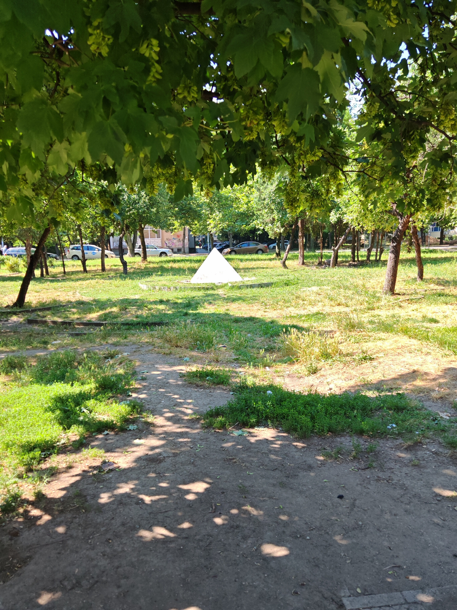 The image depicts a park-like setting with trees casting shadows on a dirt path in the foreground. In the background, there is a grassy area with a white pyramidal structure and several cars parked along a street. Buildings are visible behind the trees. The scene appears to be located in a residential area of Plovdiv, Bulgaria, as suggested by the presence of cars, buildings, and the general park aesthetic. The lighting suggests it is daytime, likely late morning or early afternoon, given the dappled sunlight. There are no people visible in the immediate foreground or engaged in any actions. The environment is characterized by lush green foliage, patchy grass, and a somewhat unkempt appearance of the park's ground cover. The white pyramidal object is the most prominent and unusual feature in the middle ground.