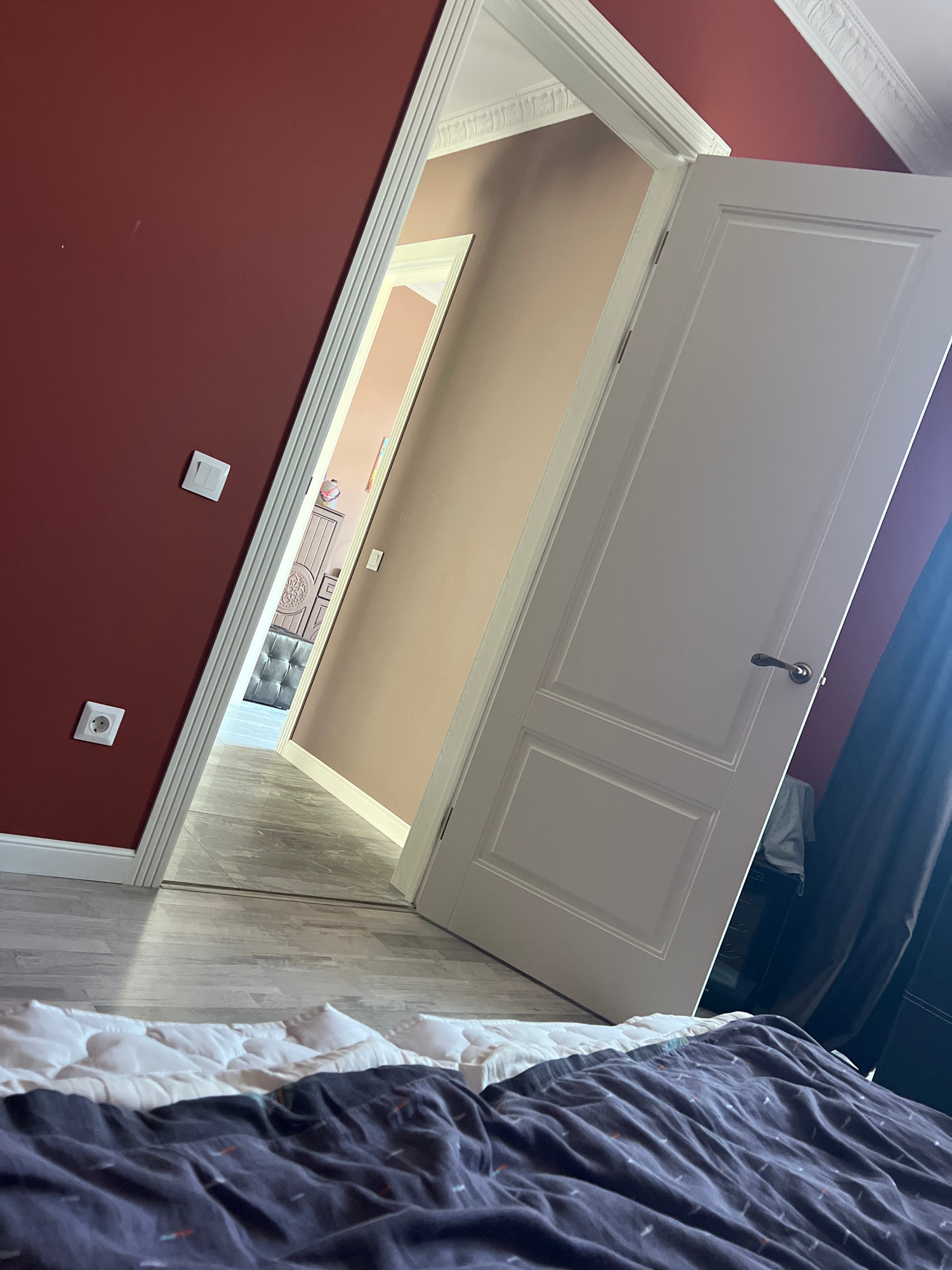 The image shows an interior view of a room, possibly a bedroom, with a dark red wall on the left and a beige wall on the right, separated by an open doorway. A white door is partially visible on the right, ajar. The floor appears to be light-colored wood or laminate. In the foreground, a bed with white bedding and a dark patterned duvet is visible. In the distance, through the doorway, a hallway leads to another room containing a bed and furniture. Light fixtures and electrical outlets are present on the walls. The scene does not provide any specific visual cues that would allow for identification of the city or a precise location within Peterhof, Russia.