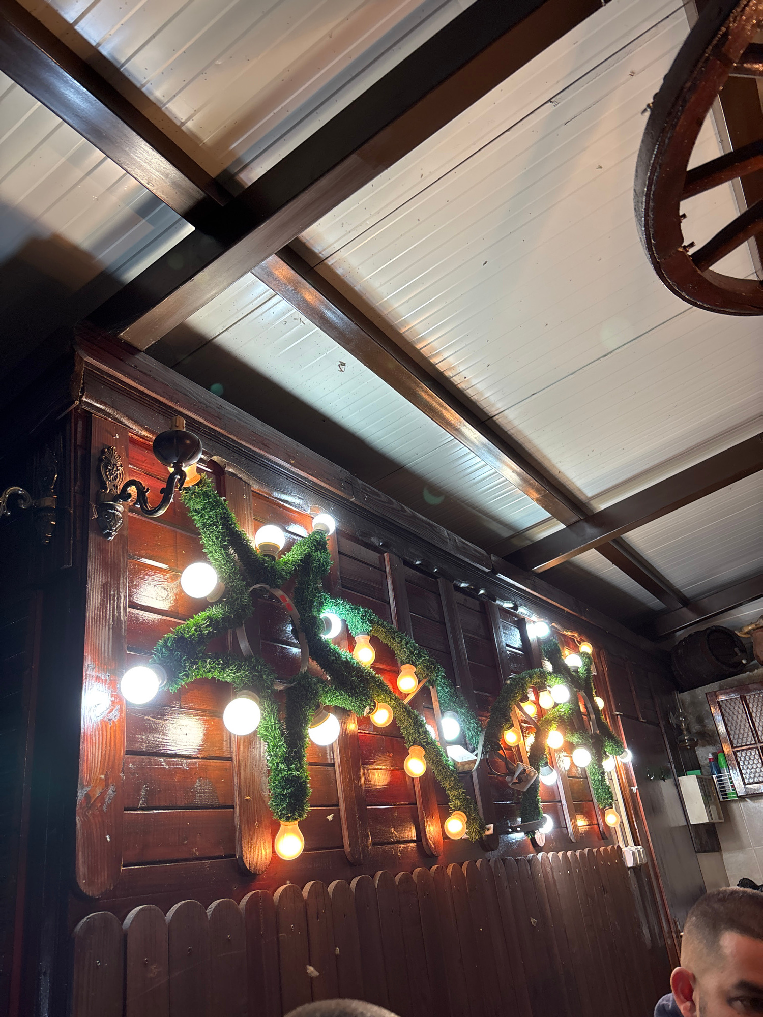 This is a low-angle, medium shot capturing a rustic, dimly lit interior space, likely an outdoor covered area or a restaurant with wooden accents. Dark brown wooden beams crisscross overhead, supporting a white, corrugated ceiling. Against a dark, varnished wooden wall, a decorative string of lights intertwined with artificial greenery is displayed. The lights are primarily round and bright white, with some emitting a warmer, yellowish glow. The greenery is lush and thick, forming a vine-like pattern across the wall. Two ornate, dark metal wall sconces are visible on the left side of the wooden wall, one of which is partially obscured by the greenery. A small, dark wooden fence runs along the bottom of the frame, creating a barrier. To the far right, a portion of a barrel and a window with a grid pattern are visible. In the foreground, the top of a person's head, with closely cropped hair, is visible in the bottom right corner, suggesting the photo was taken from a seated or low vantage point within this establishment in Zlatograd, Bulgaria.
