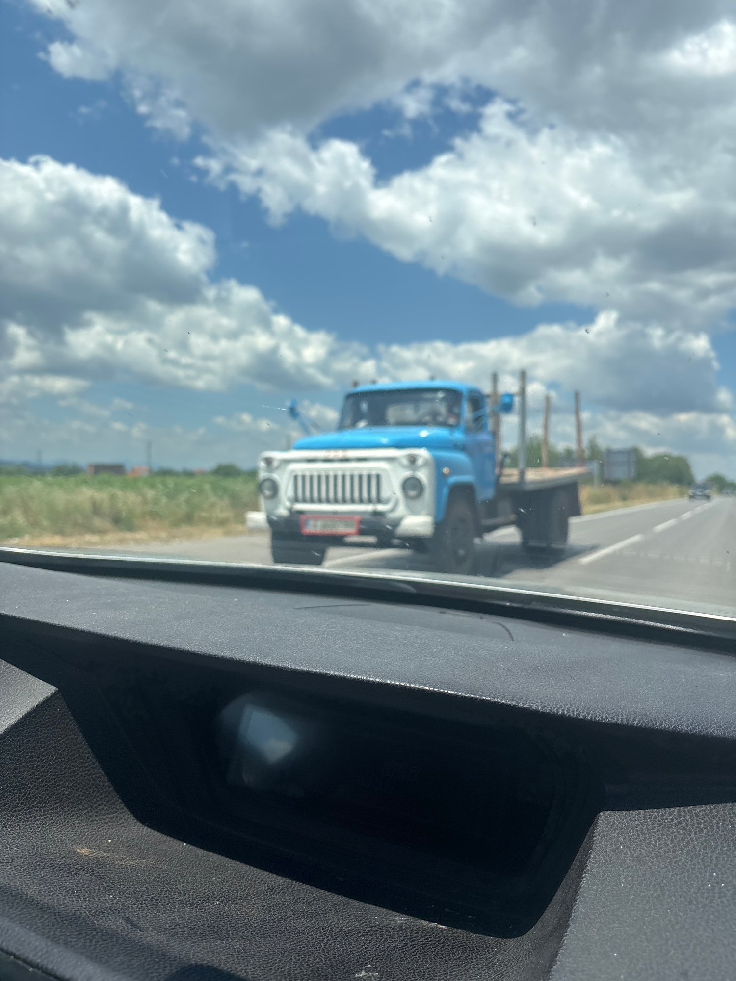 The image captures a view from inside a vehicle looking out at a bright, cloudy day with a blue sky. The foreground shows the textured interior of a car dashboard. In the middle ground, a light blue flatbed truck with a white grille and headlights is driving on a paved road. The truck appears to be loaded with wooden poles. To the left of the road, there is a grassy field with some distant buildings and trees. The road continues into the distance, where another vehicle is visible. The scene appears to be on the outskirts of the city of Manastir, Bulgaria.