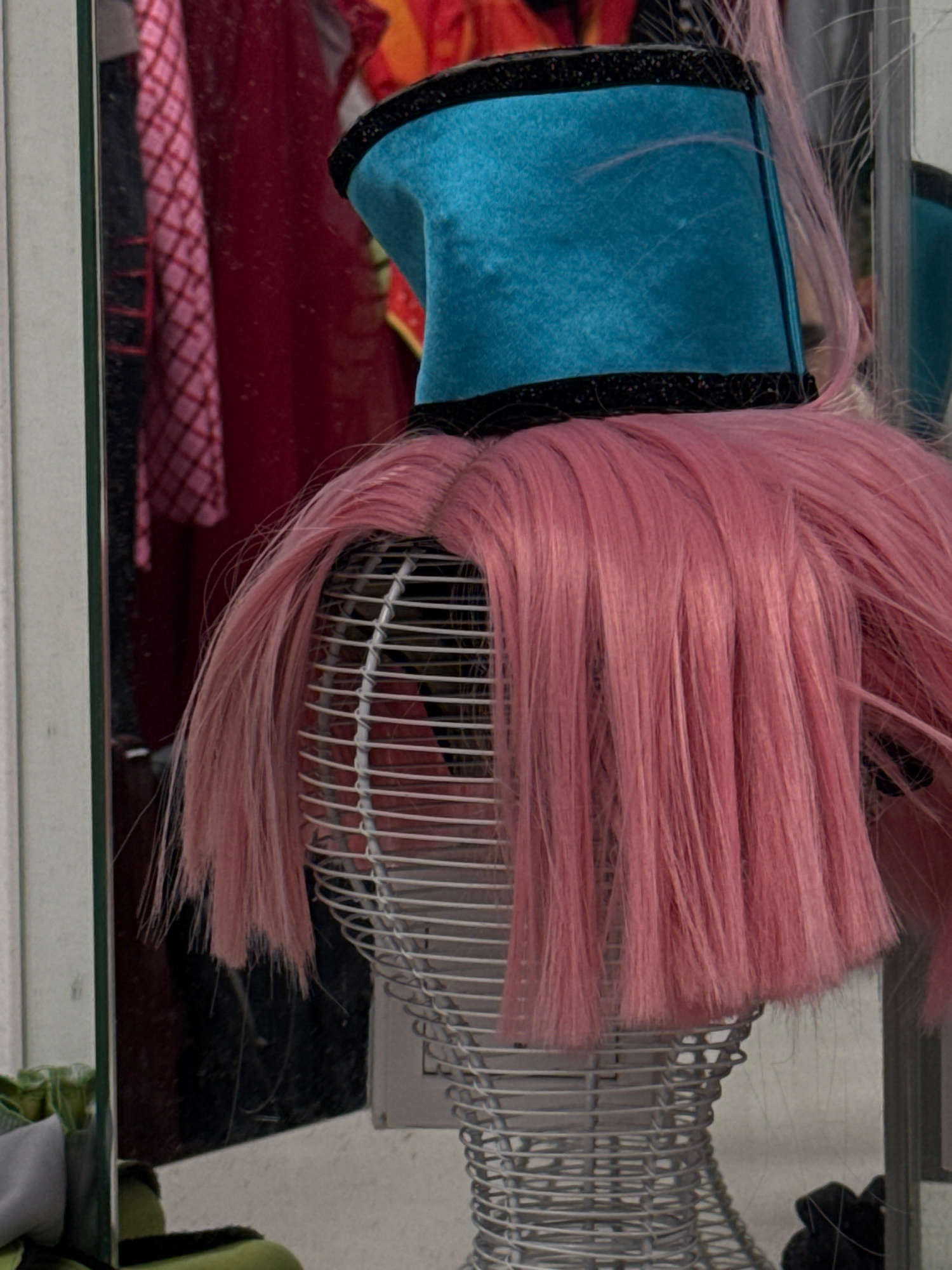The image depicts a close-up of a mannequin head covered in a vibrant pink wig. The mannequin wears a teal, cylindrical hat with black trim. The background, visible through a reflective surface, shows clothing items in red and pink. The scene appears to be indoors, possibly in a retail or display setting. There is no discernible location in Xiangzhou, China, mentioned or implied in the image.