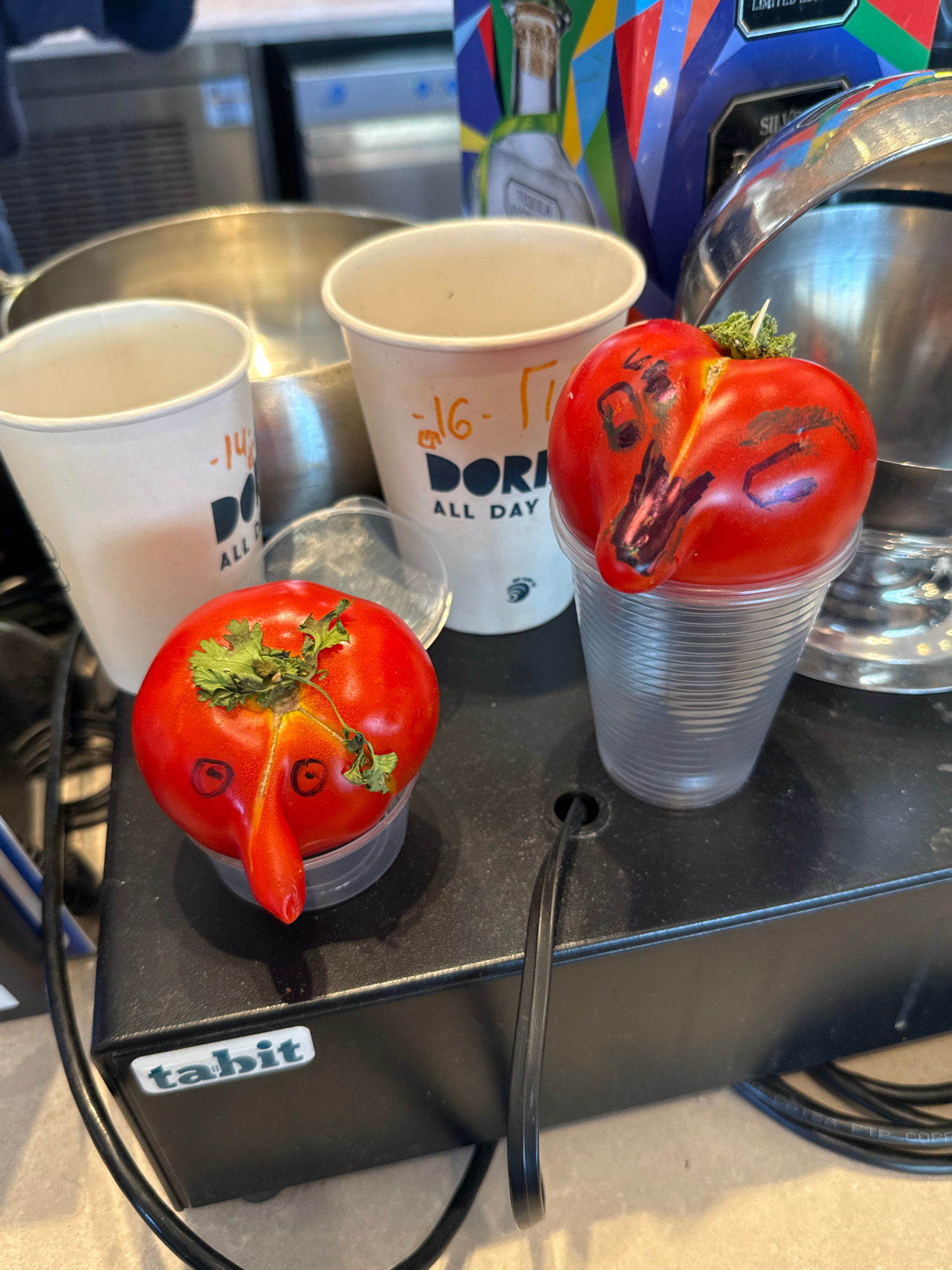 The image shows two tomatoes with faces drawn on them, placed in clear plastic cups. The tomato in the foreground has eyes drawn as black circles and a mouth as a red curved line. It also has a piece of parsley or cilantro attached to its top. The second tomato, partially visible, is also in a cup and has similar facial features drawn on it.
The tomatoes are on a black surface, which appears to be part of a machine or appliance. A black cable is visible snaking across the surface. To the left of the black surface, there are several white paper cups stacked. Some of these cups have text on them. One cup has "DOR" and "ALL DAY" written on it, along with some orange markings that look like numbers. Another cup has "ml-" written on it in orange.
Behind the cups and tomatoes, there's a glimpse of a shiny, metallic object, possibly a large bowl or container. To the far left, a colorful object with geometric patterns is visible, suggesting a vibrant background element. A sticker with the word "tabit" is affixed to the black surface.
The scene appears to be indoors, likely in a commercial setting such as a cafe or food establishment, given the presence of disposable cups and what looks like a piece of equipment. There are no people visible in the image. The lighting suggests it could be daytime. The overall impression is one of playful decoration or a creative display involving everyday objects.