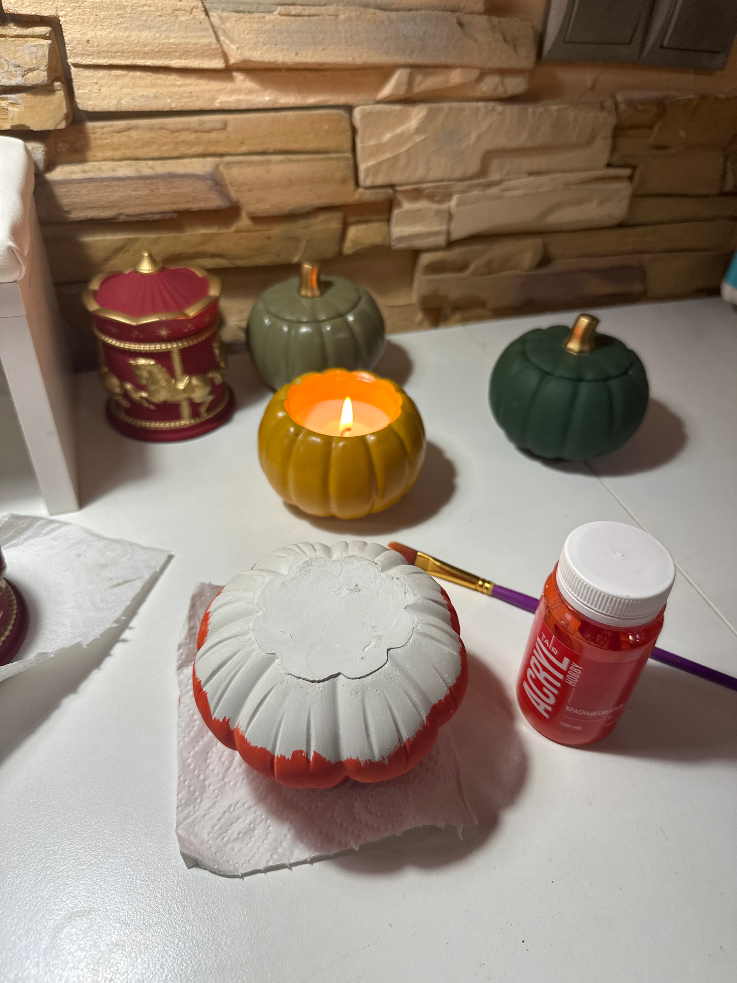 A close-up, overhead shot captures a crafting scene on a white surface, likely a table, against a backdrop of light brown, textured stone tile. In the foreground, a white pumpkin with an orange-painted base rests on a white paper towel, suggesting it is in the process of being decorated. To its right, a small bottle of red acrylic paint labeled "ACRYL HOBBY" and a purple-handled paintbrush are positioned, indicating active crafting.
Further back on the white surface, a small, lit orange pumpkin-shaped candle casts a warm glow. To its left are two decorative, dark green pumpkins with gold stems, and behind them is a red, cylindrical trinket box adorned with gold detailing, including a horse-and-rider motif.
The scene appears to be indoors, possibly in a home or workshop, with no visible indications of its specific location within Vladivostok, Russia. The lighting is artificial, highlighting the objects and textures present.