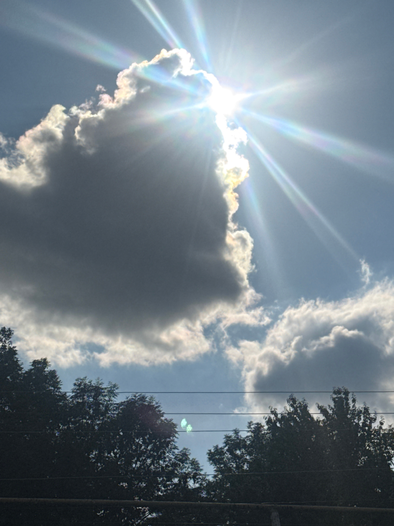 The image captures a bright, sunlit sky with dramatic clouds. The sun is a prominent feature, positioned in the upper left quadrant, emitting strong rays that create a starburst effect. These rays refract and scatter through the atmosphere, producing subtle rainbow-like hues.
Several large, dark gray clouds dominate the center of the frame, with the sun partially obscured by one of them. The edges of these clouds are illuminated by the sun, creating a stark contrast. Lighter, wispy clouds are visible higher up in the sky, suggesting a dynamic weather pattern.
On the right side of the image, silhouetted trees with dense foliage are visible. Their dark forms stand out against the bright sky. Several parallel utility lines run vertically through the scene, crossing in front of the trees and the sky.
The scene appears to be outdoors, under a daytime sky. The bright sunlight suggests it is mid-day or afternoon. The presence of trees and utility lines indicates a potentially rural or suburban setting. The overall mood is one of bright natural light and perhaps a slightly dramatic or awe-inspiring sky. There is no visible text in the image.