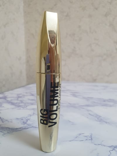 A gold-colored mascara tube stands upright on a marble-patterned surface. The tube has black text that reads "BIG VOLUME EXPLOSION" vertically along its side. The background is a textured, light-colored wall. The image is a still shot, with no people or actions depicted. The environment appears to be an indoor setting. The location in Tashkent, Uzbekistan, is not discernible from the image itself.