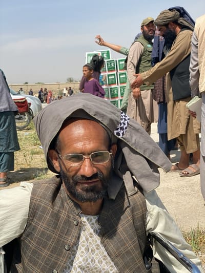 The image shows a middle-aged man wearing a gray head covering, glasses, and a patterned brown vest, seated in what appears to be a wheelchair. The background includes a crowd of people, some wearing traditional Afghan clothing, and stacks of green boxes with white writing. The location is likely Kandahar, Afghanistan, based on the attire and environment. A boy stands near the boxes, and several men are engaged in what looks like a greeting or exchange. A motorcycle is visible in the background. The overall scene suggests a public gathering, possibly related to aid distribution.
