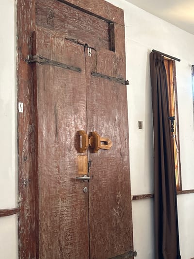 A tall, weathered, double wooden door with a dark reddish-brown finish dominates the left side of the image. The wood grain is highly pronounced, with visible wear and tear, and lighter colored streaks where the finish has worn away. There are several dark, metal hinges attached to the door. The left door has a small white electrical outlet cover with what appears to be a light switch on it. The right door has a silver doorknob and a keyhole. In the center of the door, there are two square, light-colored wooden latch mechanisms.
To the right of the door, a plain white wall extends upwards. Partially visible on the wall are two small, square, dark-colored wall plates. Further to the right, a dark brown curtain is drawn across a window that has a wooden frame and thin, vertical wooden slats. The bottom of the curtain reaches down to a horizontal wooden beam attached to the wall.
The scene appears to be an interior space, possibly a hallway or entrance. The overall impression is one of age and rustic character. The location is stated as North Kuta, Indonesia.