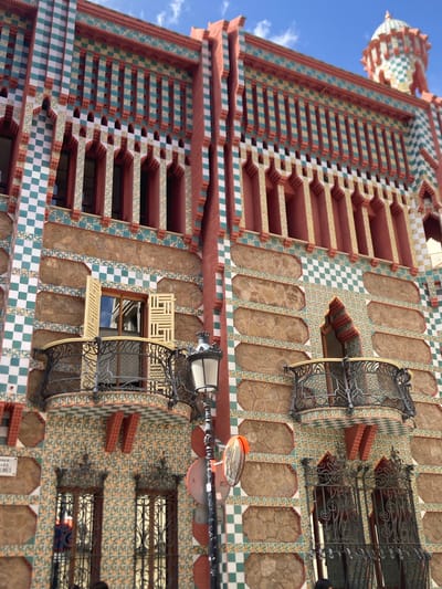 The image shows the exterior of a building with a highly decorative facade. The building is located in Barcelona, Spain. The facade features a vibrant pattern of green and white checkerboard tiles, red brickwork, and stone panels. Several windows are visible, some with ornate wrought-iron balconies. One window has light-colored shutters that are open. A street lamp with a reflective traffic mirror is positioned in the foreground, slightly to the right of the center. The sky above is blue with a few wispy clouds.